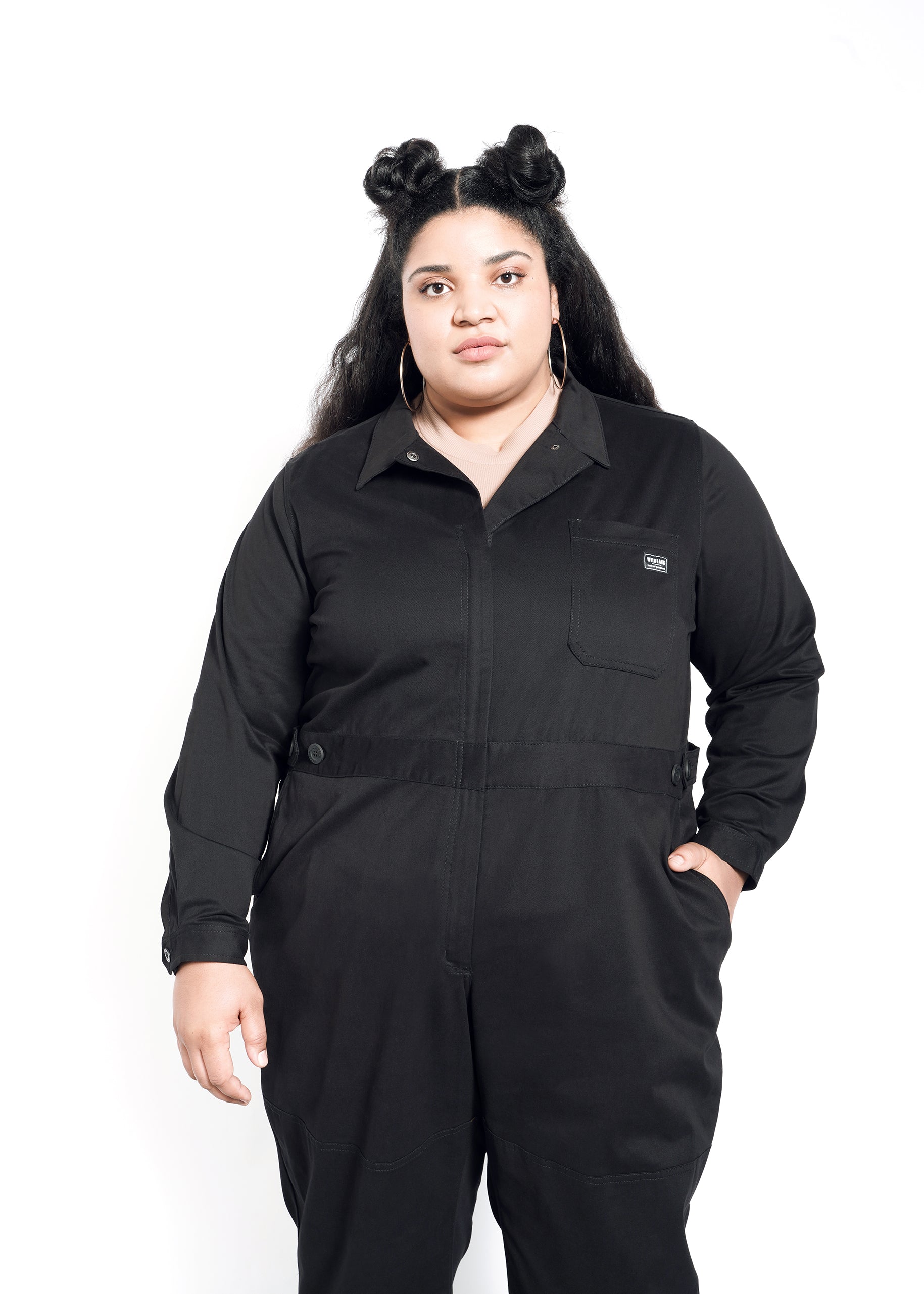 A person with dark hair in two buns confidently wears The Essential Long Sleeve High Waisted Coverall, made of cotton stretch with an adjustable waist, hands in pockets, and hoop earrings, standing against a white background.