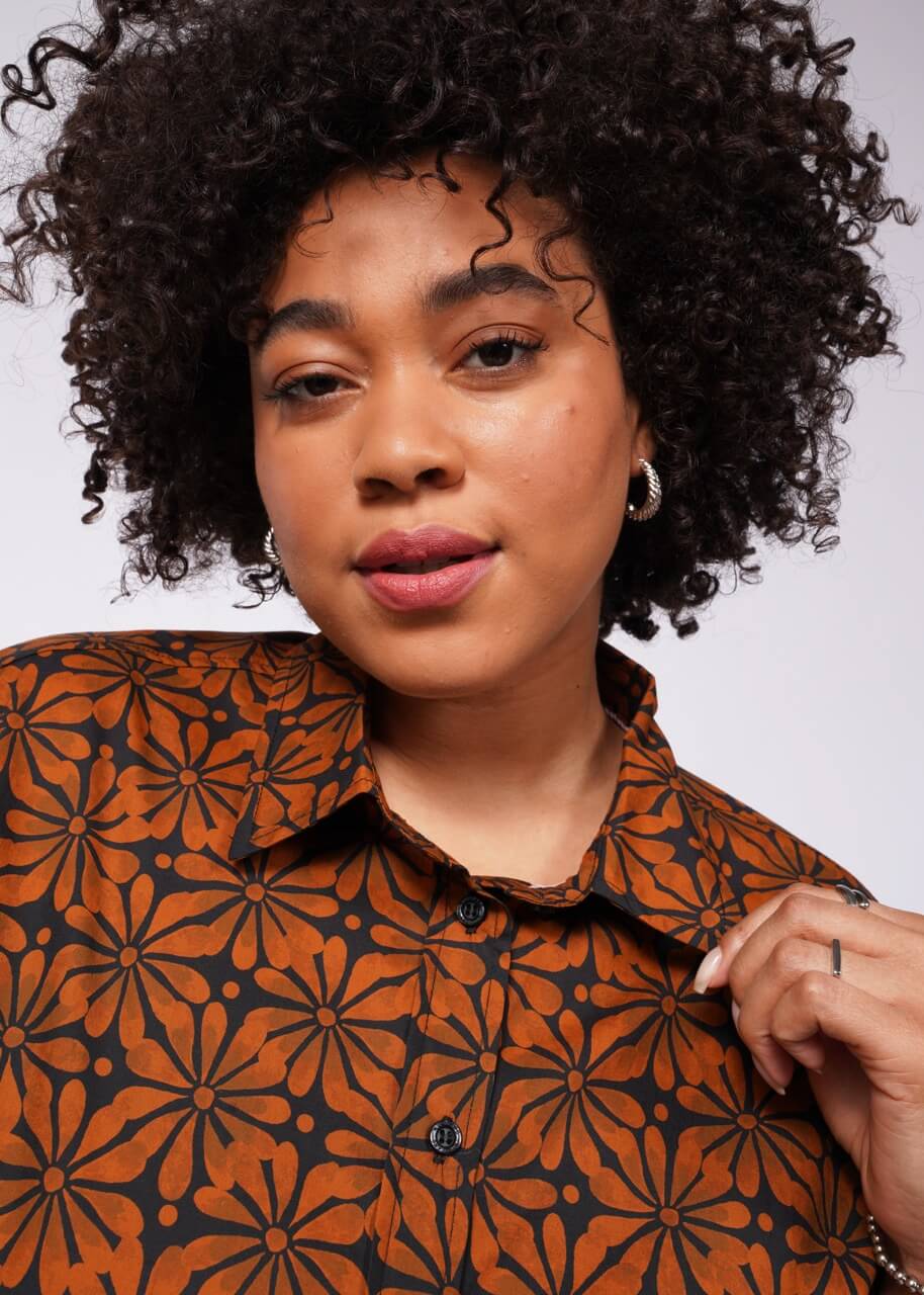 A person with curly hair models The Essential Button Up, a tailored fit brown shirt featuring a black floral pattern, as they look into the camera. They wear small hoop earrings and hold the collar in one hand. The breathable cotton fabric drapes perfectly against a plain, well-lit background.