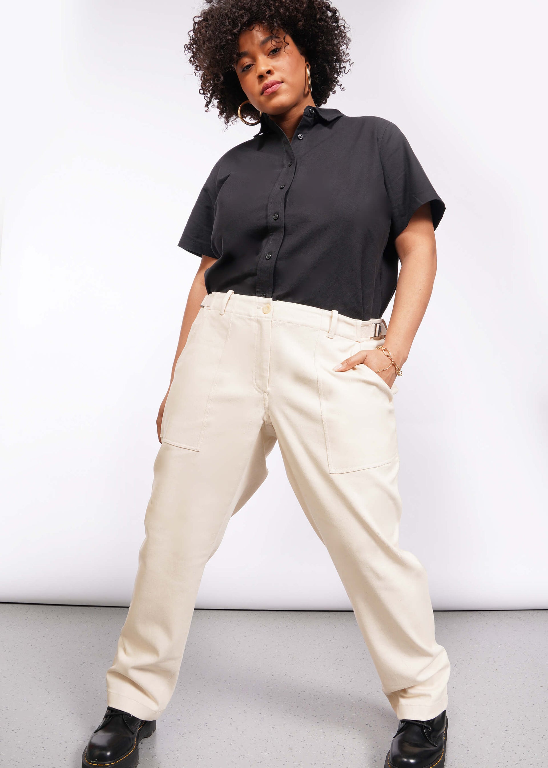 A person with curly hair and hoop earrings poses confidently in a black short-sleeved button-up shirt and light-colored, adjustable waist Essential Denim Utility Pant against a plain white background.