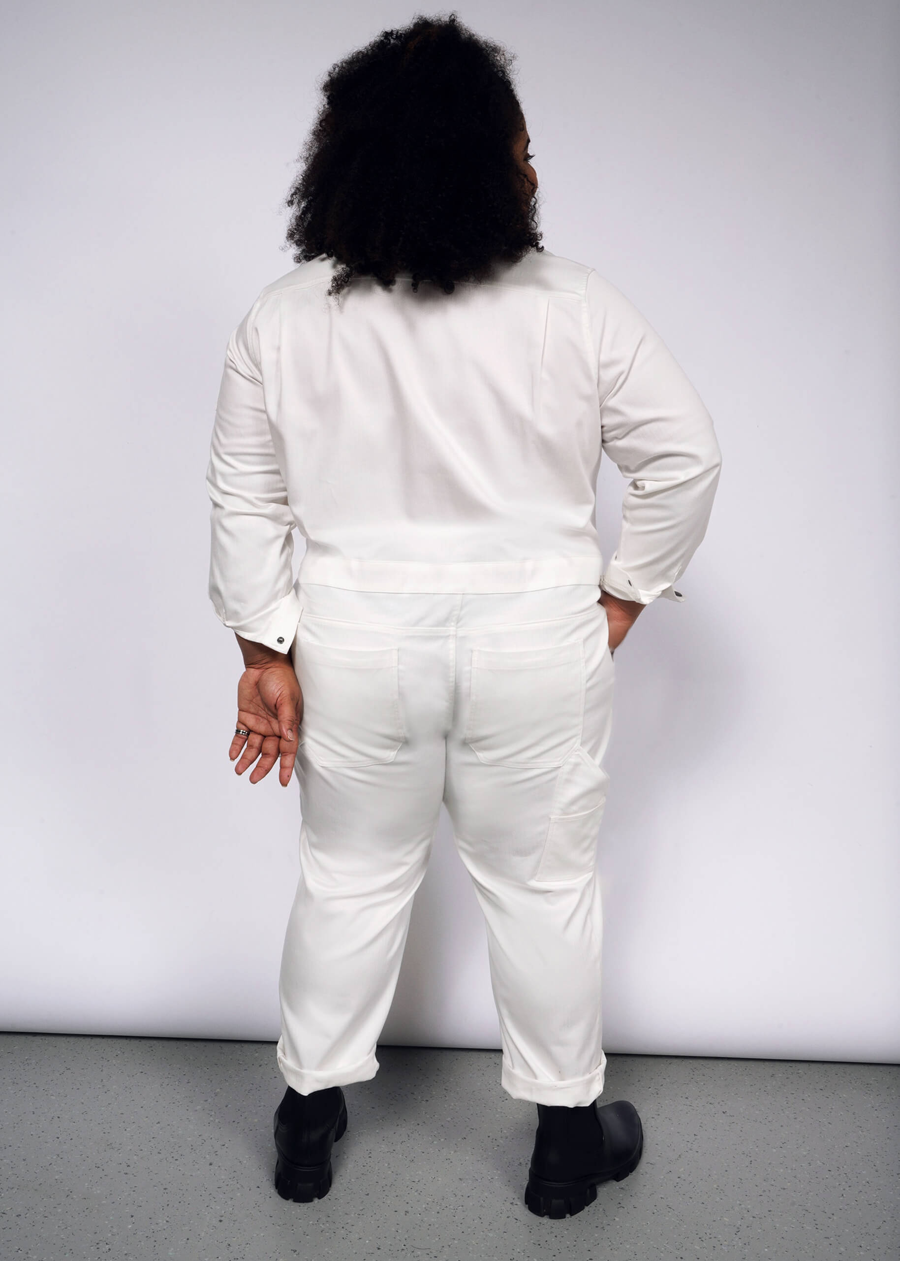 A person with curly hair is seen from behind, wearing The Essential Long Sleeve Herringbone Coverall in white, featuring a full-length zipper. They pair it with black shoes and stand against a plain white backdrop, holding a small object in their right hand.