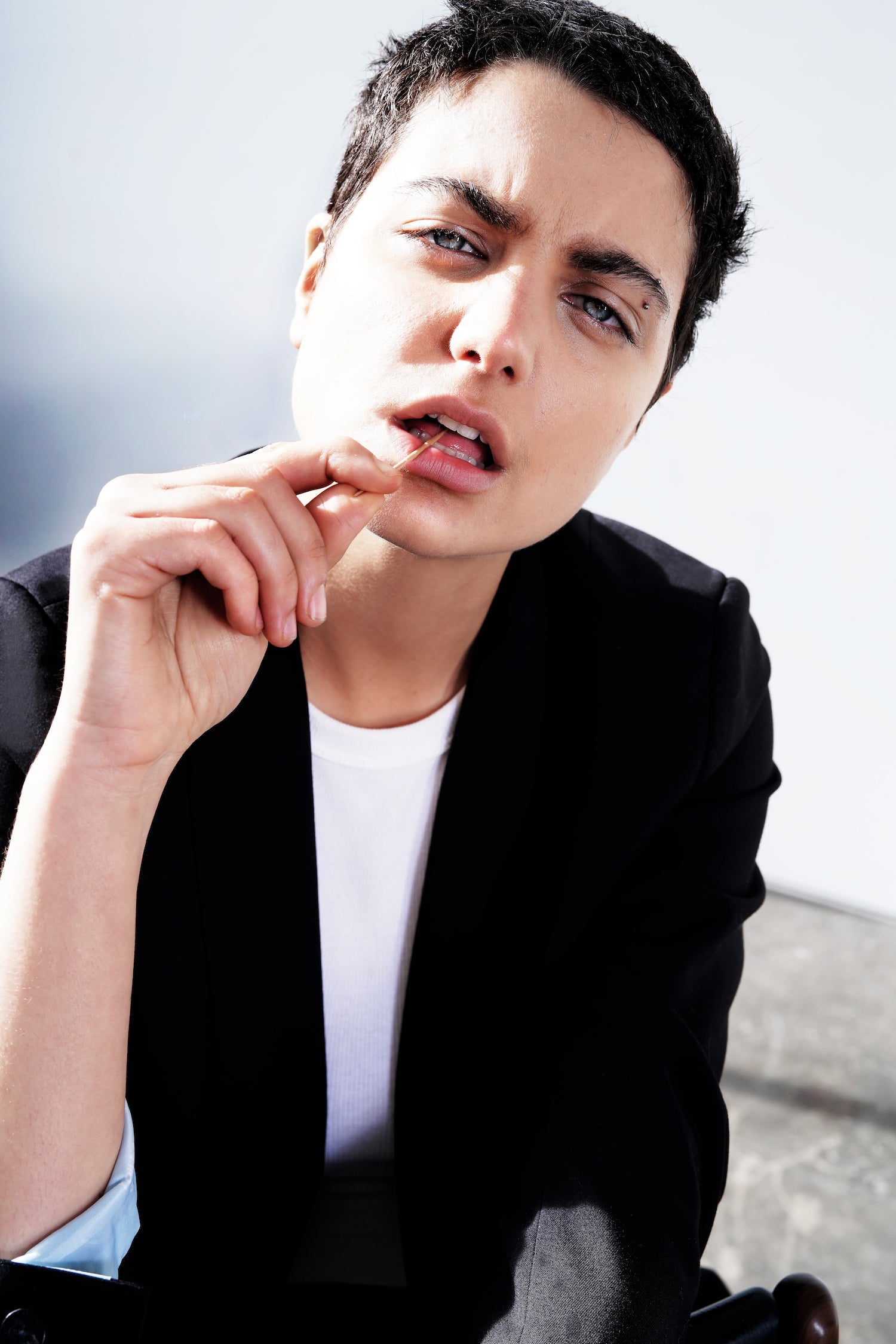 Person with very short black hair and wearing black blazer looks at camera while holding toothpick in mouth.