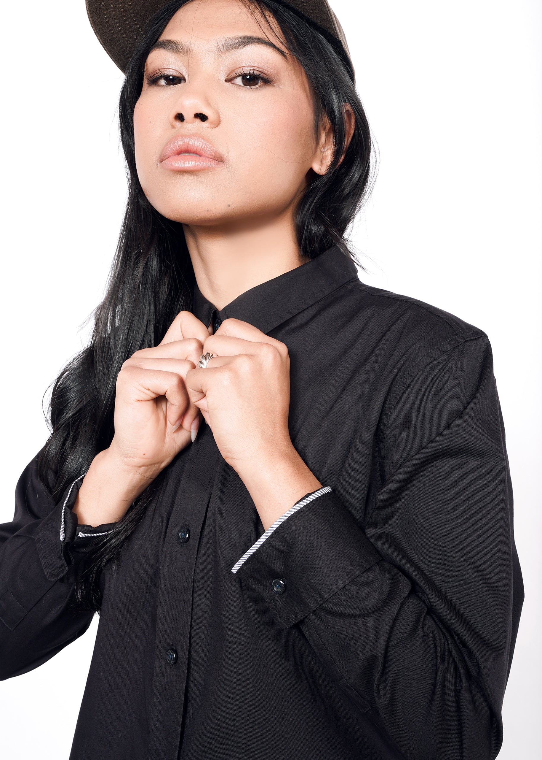 A person with long black hair confidently adjusts their collar, wearing The Essential Long Sleeve Oxford Button Up and a cap, against a white background.