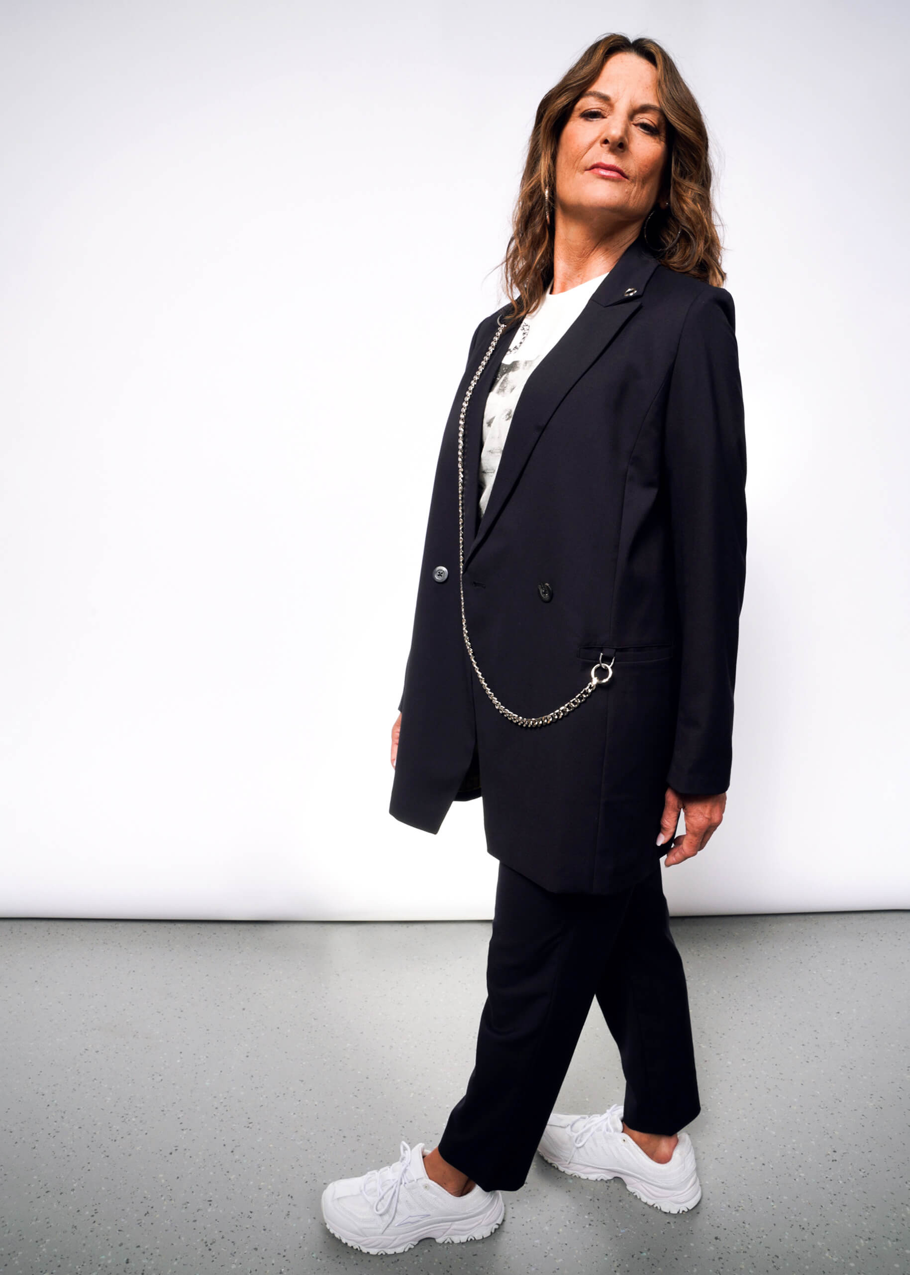 A person confidently poses in the Debbie Harry X Wildfang Double Breasted Blazer with metal detailing and a chain accessory. They have long brown hair, a white shirt, and sneakers, against a plain white background, slightly angled to the side.