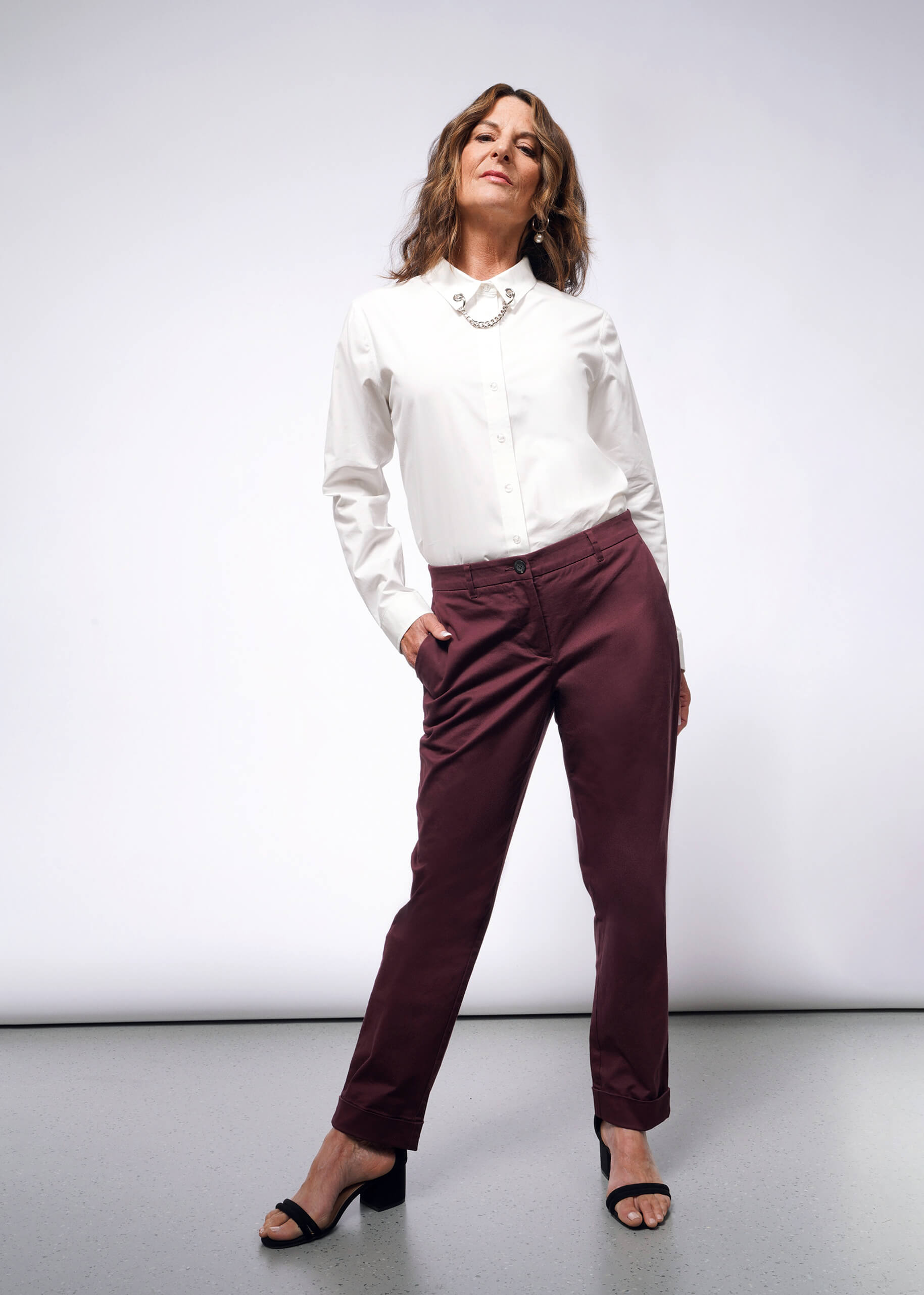 A woman stands confidently against a plain background, wearing a white button-up shirt with convertible cuffs, The Essential Trouser in maroon—a wardrobe staple—and black sandals. Her hands rest in her pockets while adorned with a necklace, her expression self-assured.