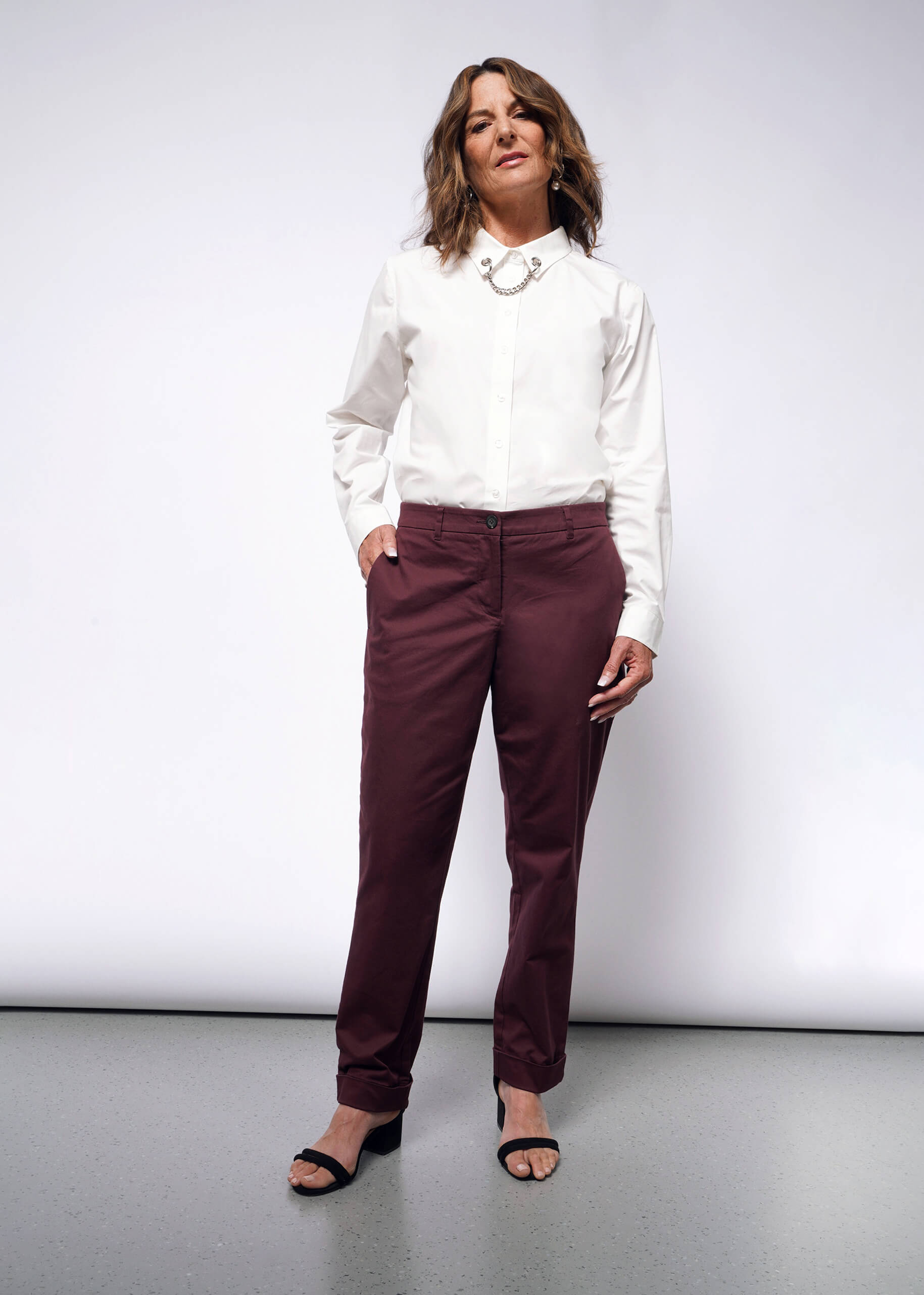 Against a plain background stands a person with shoulder-length brown hair. They exhibit confidence in a white button-up shirt with convertible cuffs, The Essential Trouser in burgundy, and black open-toed heels. A chunky silver necklace completes the ensemble.