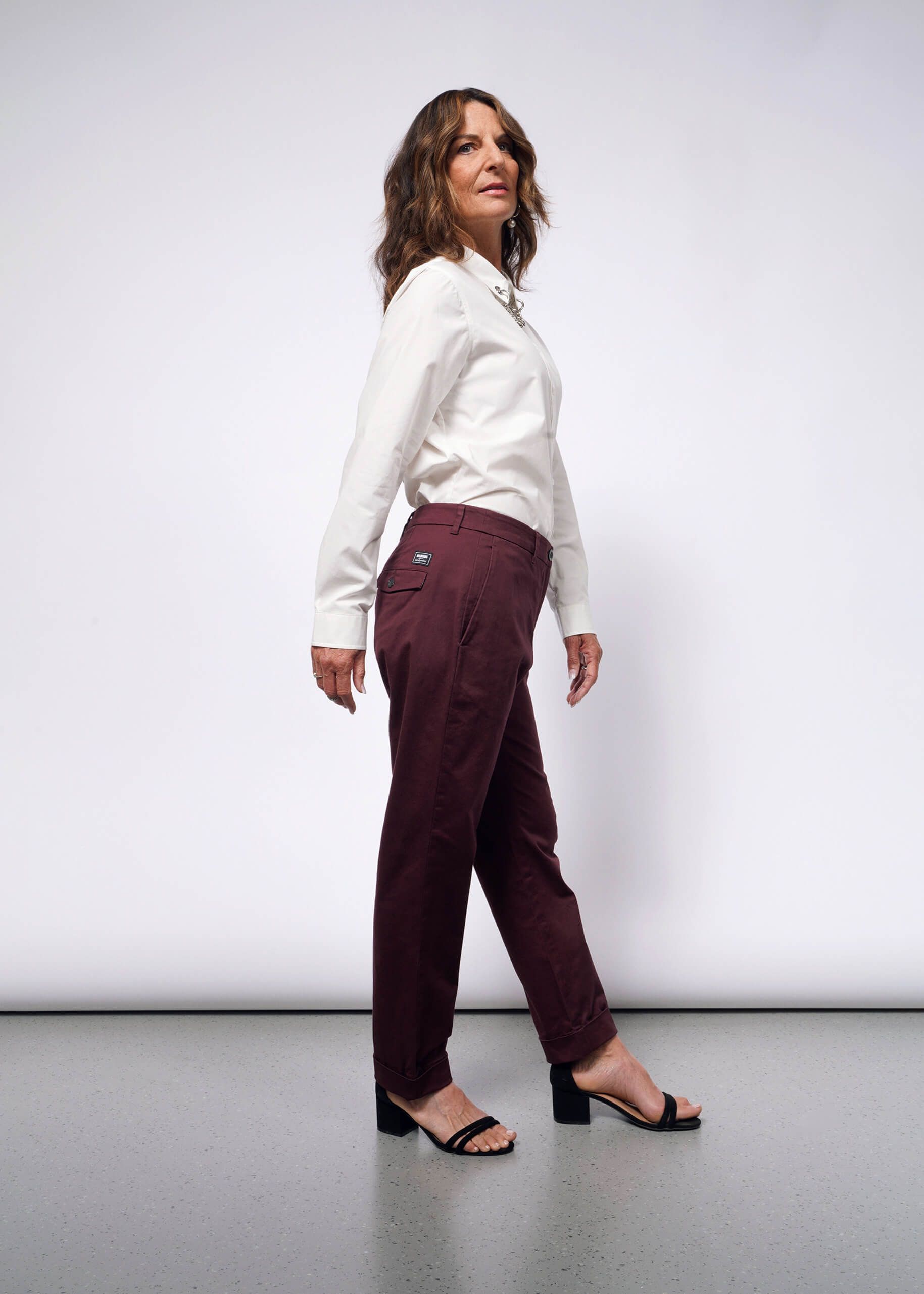 A person with long hair poses sideways in a white long-sleeve shirt with convertible cuffs and The Essential Trouser in maroon, a wardrobe staple. They complete the look with black sandals against a plain white background.