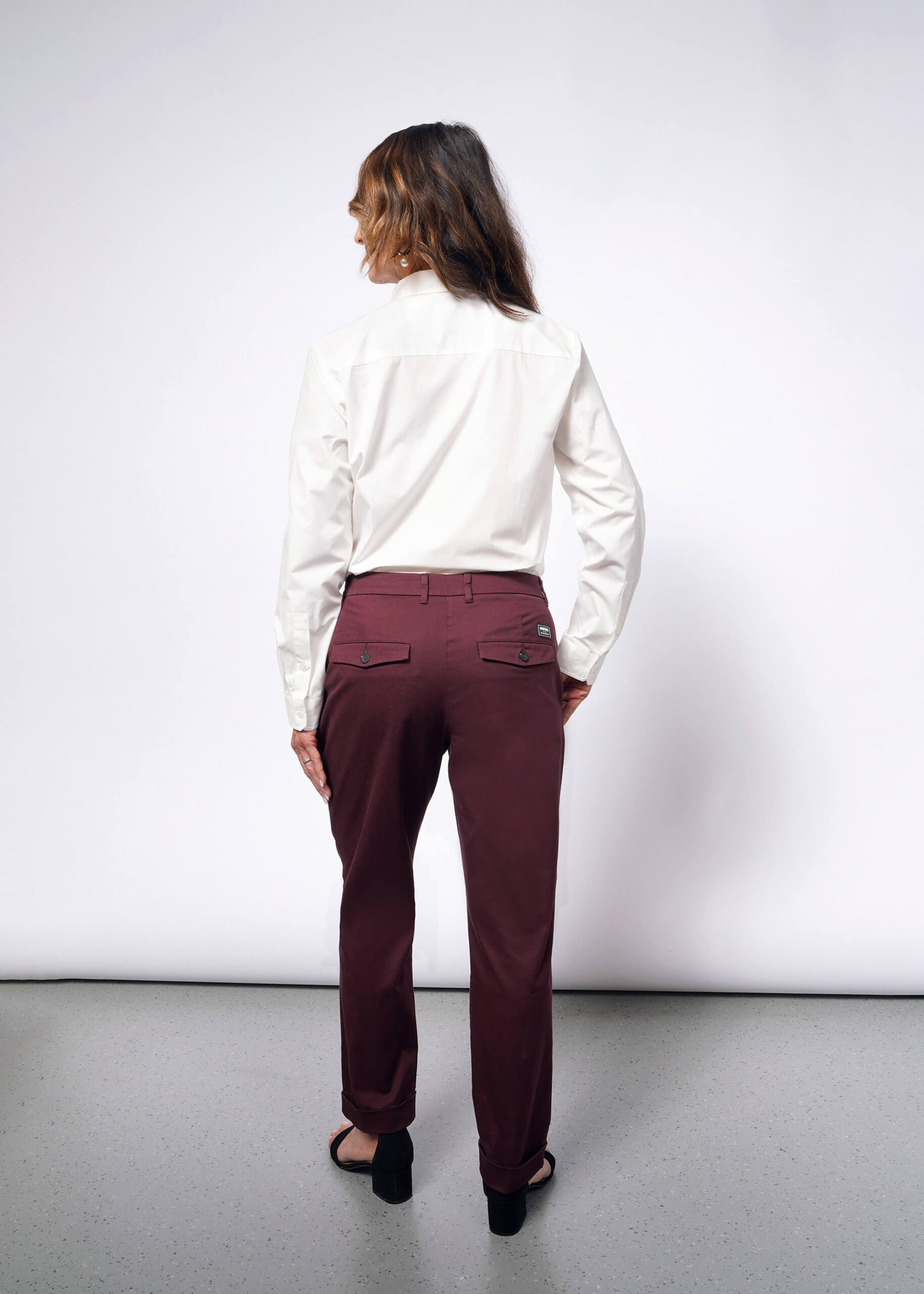 Facing away from the camera, a person confidently models maroon The Essential Trouser, paired with a white long-sleeve shirt featuring convertible cuffs and black heeled sandals. Their long, wavy hair adds to the stylish look against a plain white background.