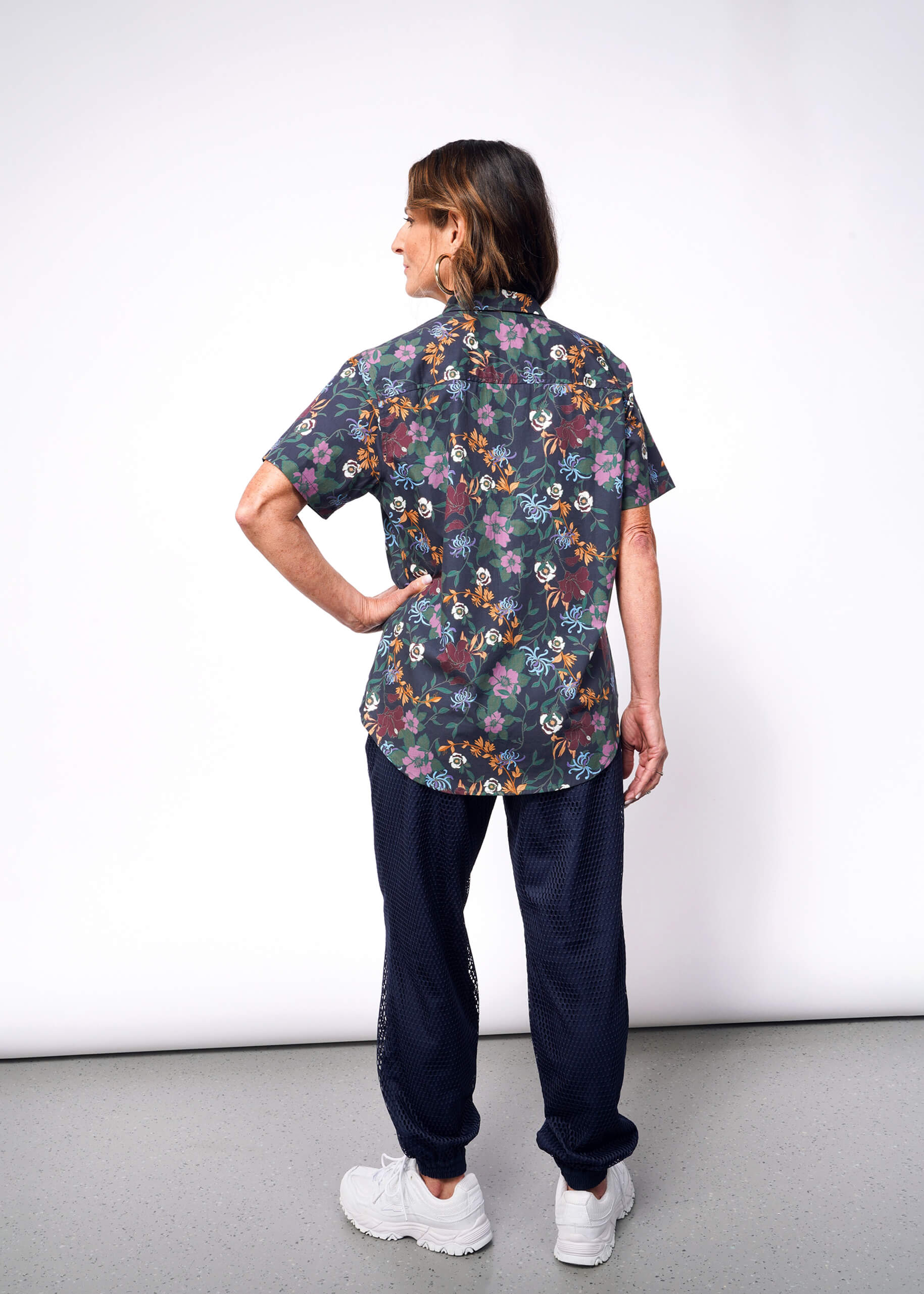 A person with shoulder-length brown hair shows off The Essential Button Up, featuring a dark floral Wildfang print, paired with tailored navy pants and white sneakers. They rest one hand on their hip against a plain light gray background.