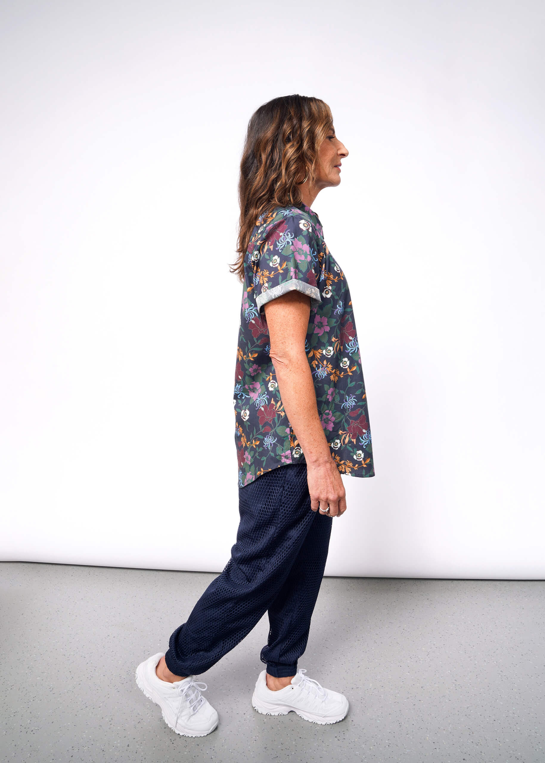 A person with long hair, wearing The Essential Button Up in a floral Wildfang print made of 100% cotton, dark tailored-fit pants, and white sneakers, stands in profile against a plain white background.