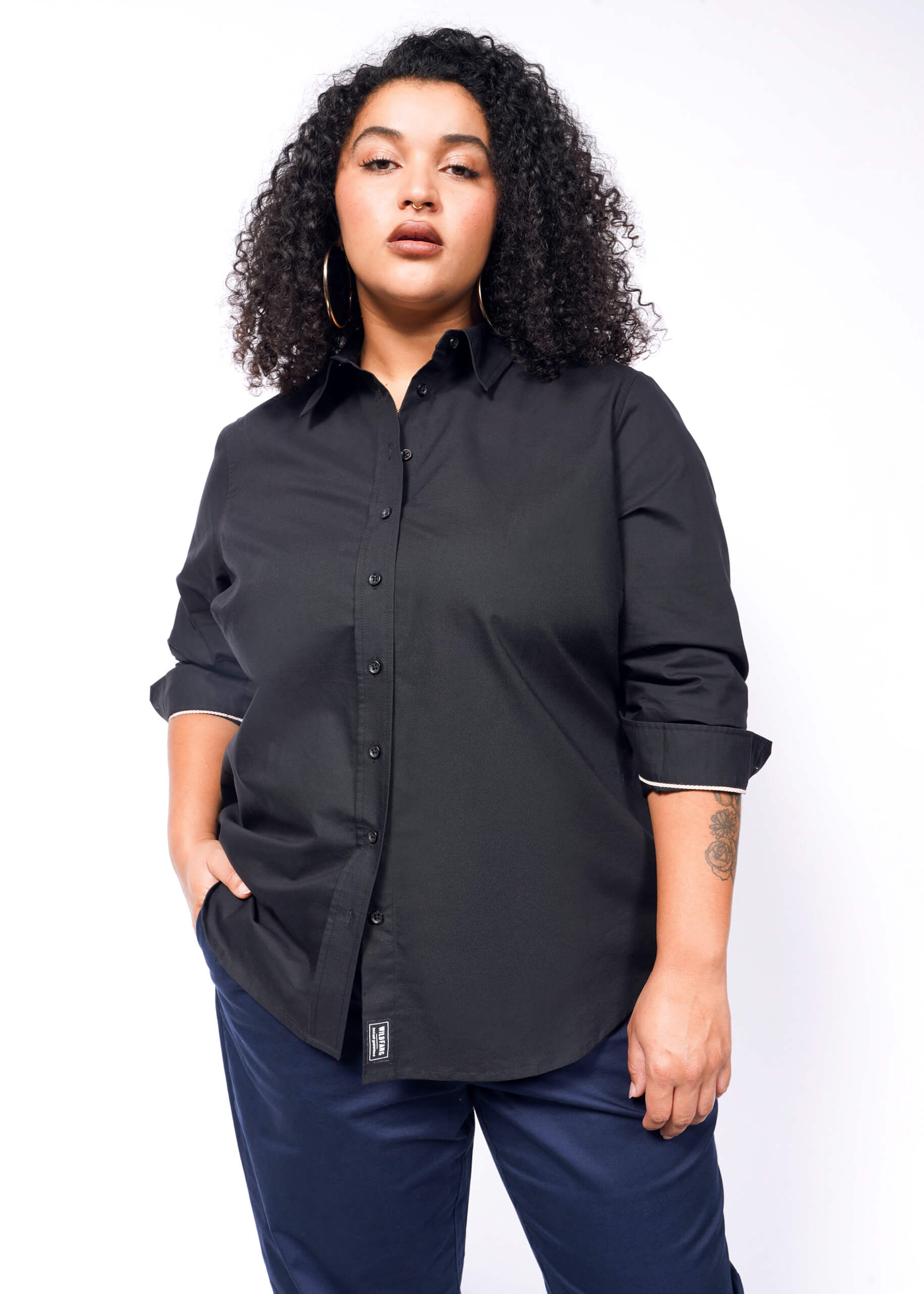 A curly-haired person in The Essential Long Sleeve Oxford Button Up, a tailored fit, black, 100% cotton shirt, pairs it with dark pants and stands with one hand in their pocket.