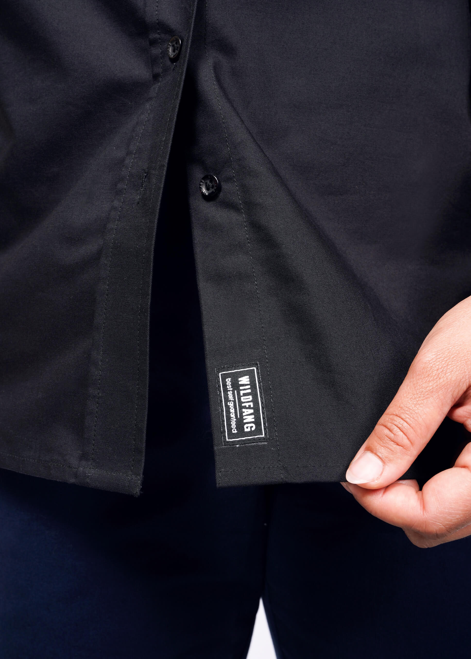 A person lifts the hem of The Essential Long Sleeve Oxford Button Up, a black, tailored-fit shirt, exposing a small WILDFANG label in white. This 100% cotton shirt is unbuttoned at the bottom and elegantly paired with dark blue pants.