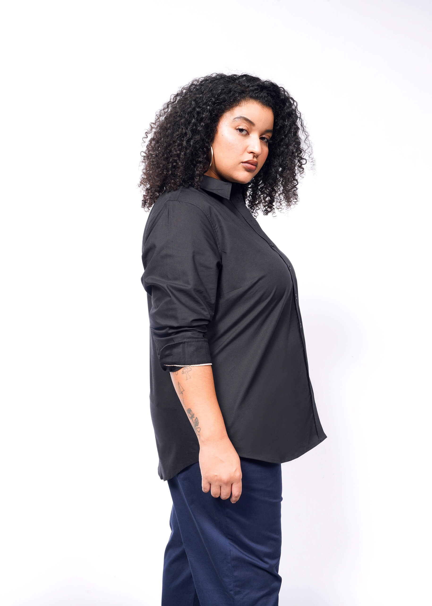 Against a plain white background, a person with curly hair stands in profile. They wear The Essential Long Sleeve Oxford Button Up in black, with rolled-up sleeves and perfectly tailored blue pants, exuding a confident expression.