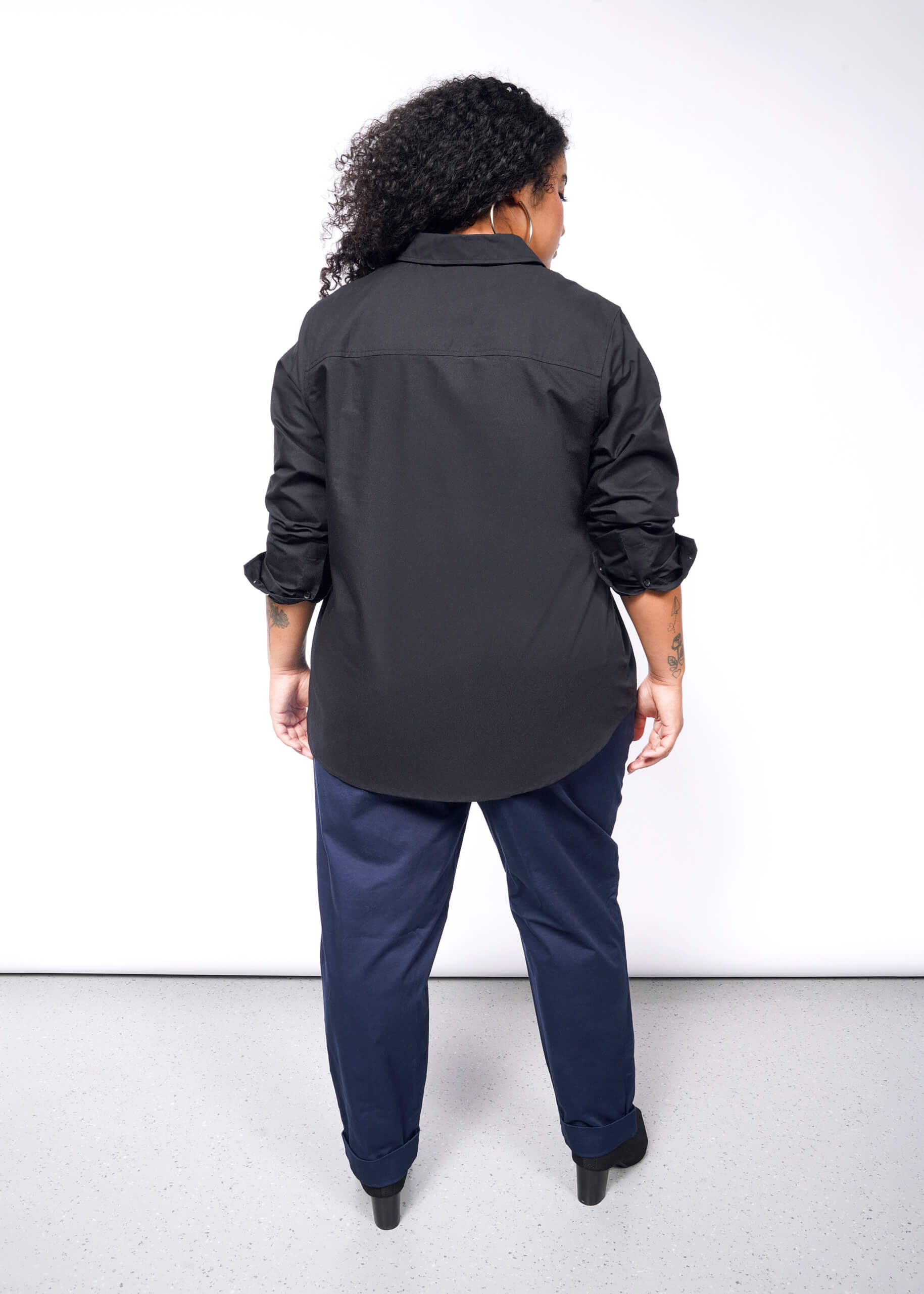 A person with curly hair stands against a white background, wearing The Essential Long Sleeve Oxford Button Up with rolled-up sleeves, navy blue pants, and black shoes. Their tattooed forearms add a distinctive touch to the look.