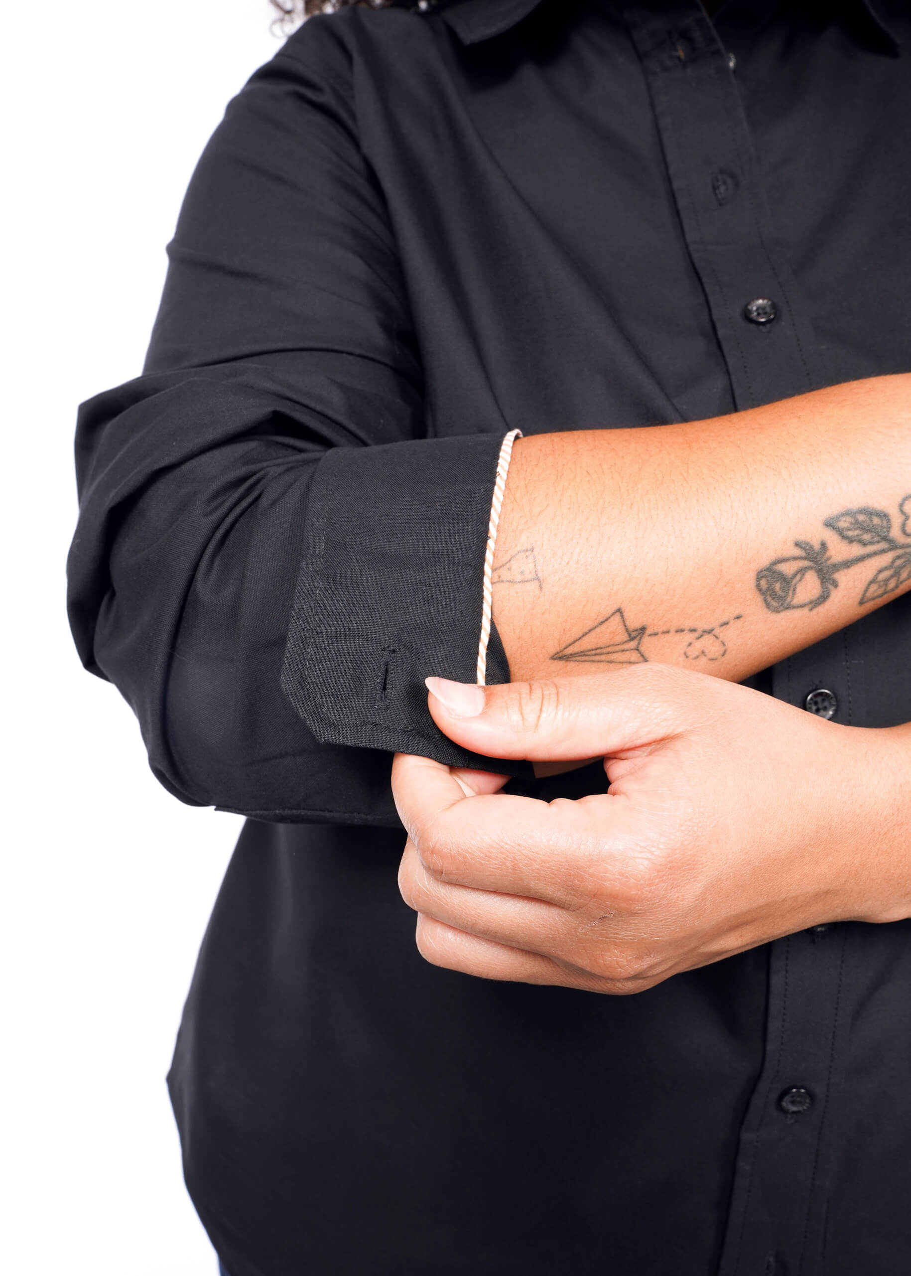 Wearing The Essential Long Sleeve Oxford Button Up, a 100% cotton tailored-fit shirt, a person rolls up their sleeves, exposing forearm tattoos of a paper plane, heart, and rose, while using their right hand to adjust them.