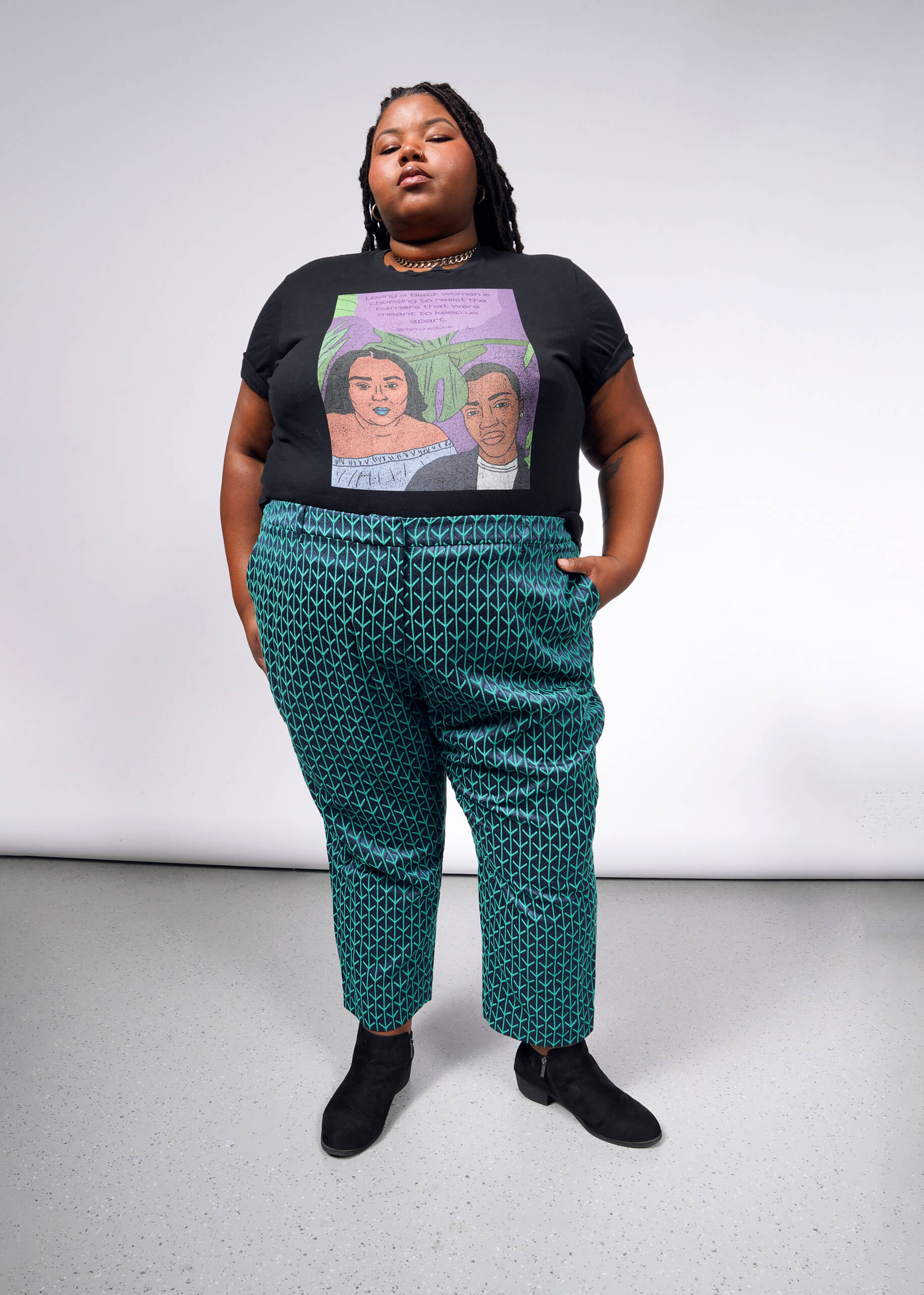 Against a light background, a confident individual wears The Empower Jacquard Slim Crop Pant in green and a black T-shirt with a colorful design. Theyre styled with black boots, a necklace, have long braided hair with one hand in their pocket.