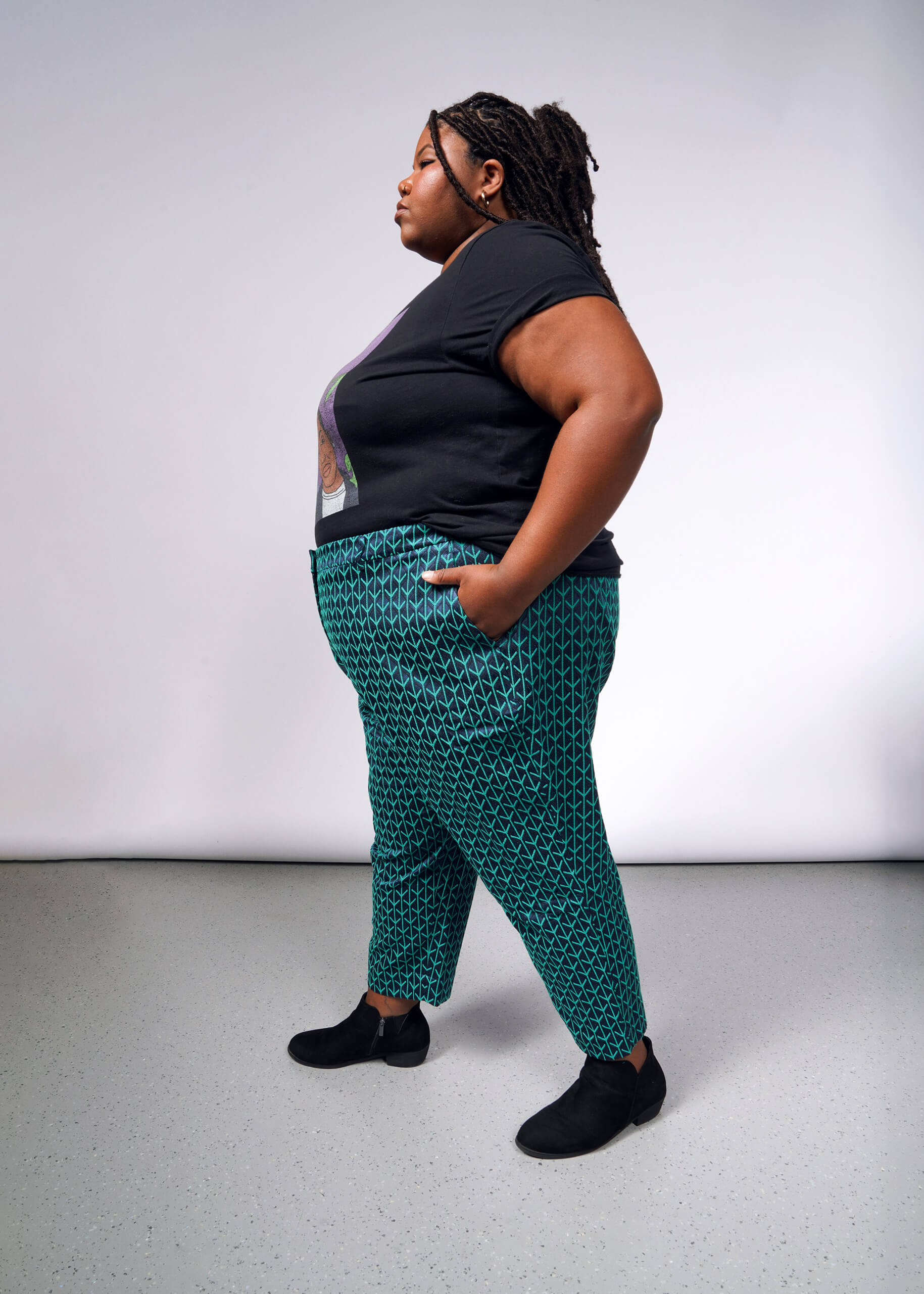 A person with long braided hair stands in profile against a plain backdrop, wearing a black T-shirt, The Empower Jacquard Slim Crop Pant in patterned blue-green jacquard fabric with a high-rise fit, and black shoes. Their hands are in pockets, conveying a confident expression.