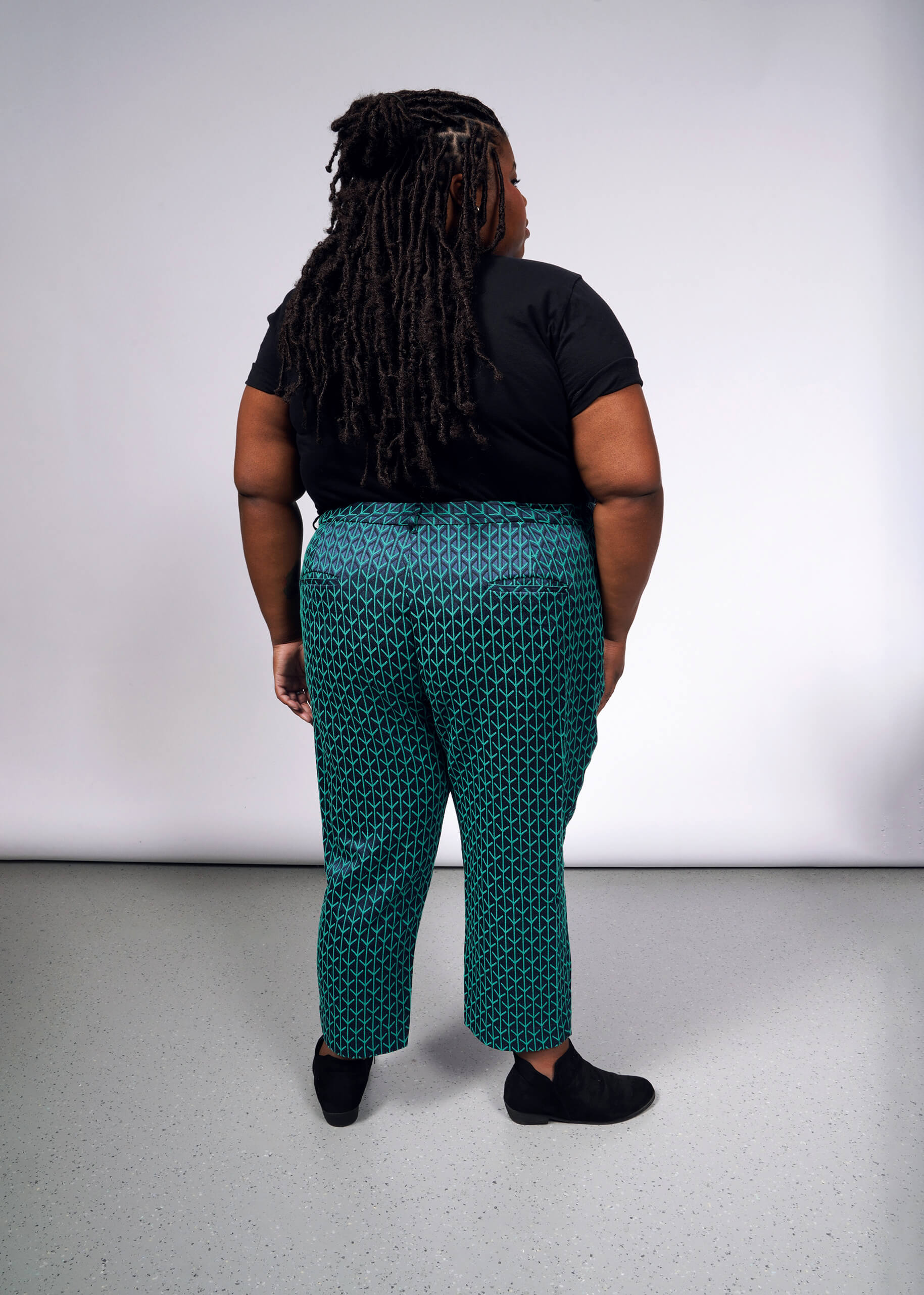 A long-haired person faces away, wearing a black t-shirt, The Empower Jacquard Slim Crop Pant in teal with a high-rise fit, and black shoes on a light gray floor against a white backdrop.