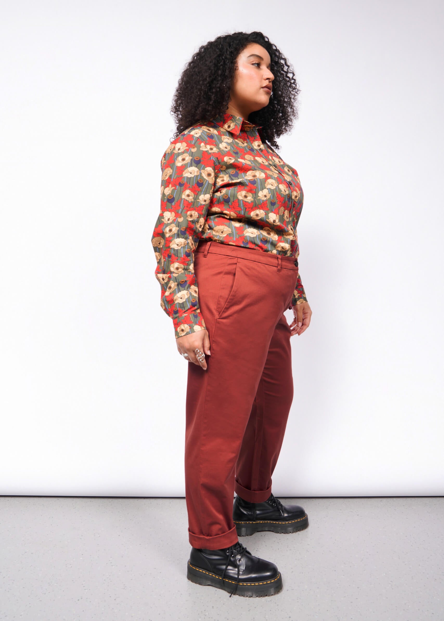 With their curly hair, a person confidently wears The Essential Trouser featuring a relaxed mid-rise fit. A colorful floral blouse tops the wardrobe staple, paired with black platform shoes for an edgy touch. The plain white background highlights their stylish ensemble.