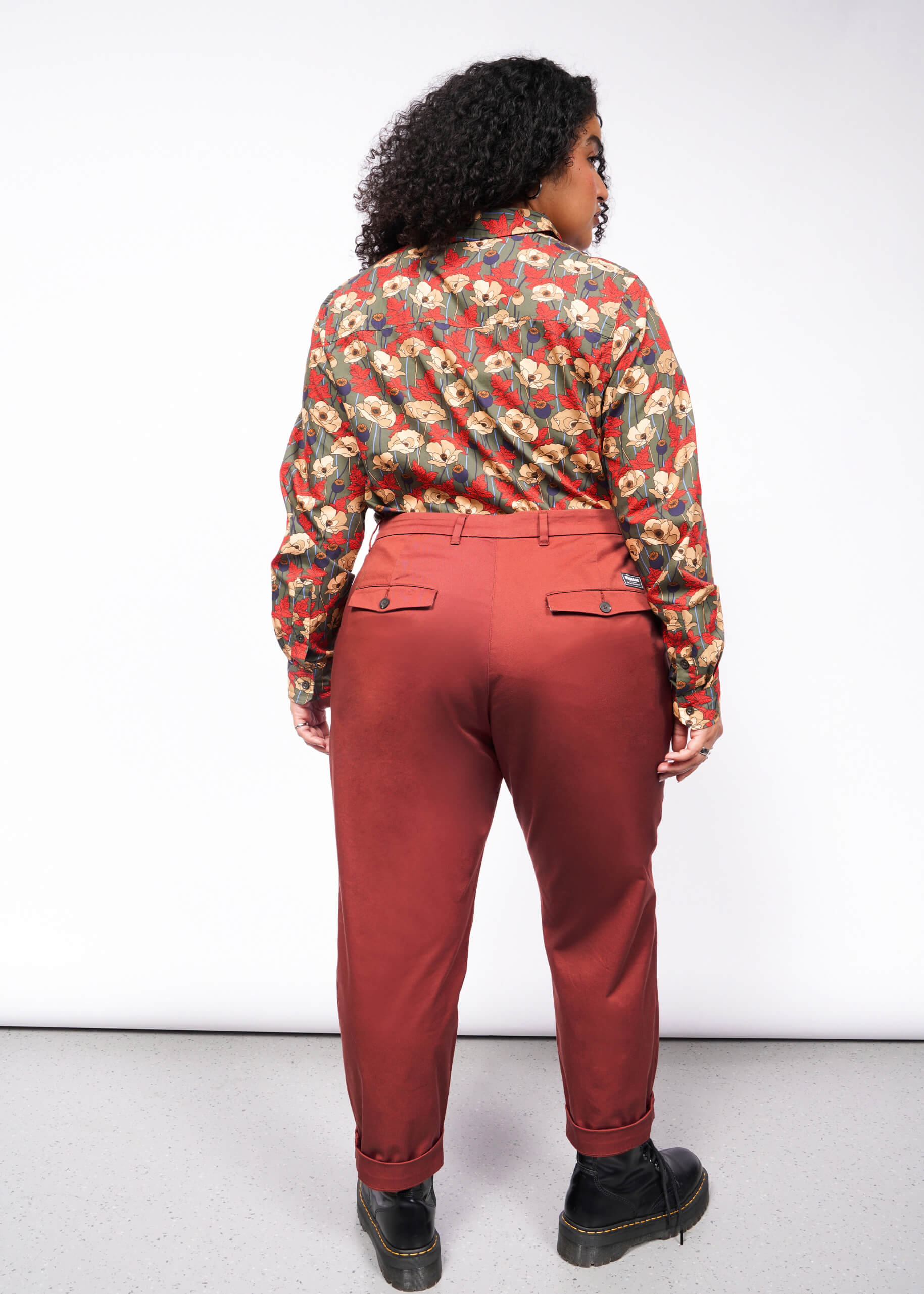 A person with curly hair stands with their back to the camera, showcasing The Essential Trouser in a classic rust color and relaxed mid-rise fit, paired with a floral shirt. Black shoes complete this timeless ensemble against a plain white backdrop.