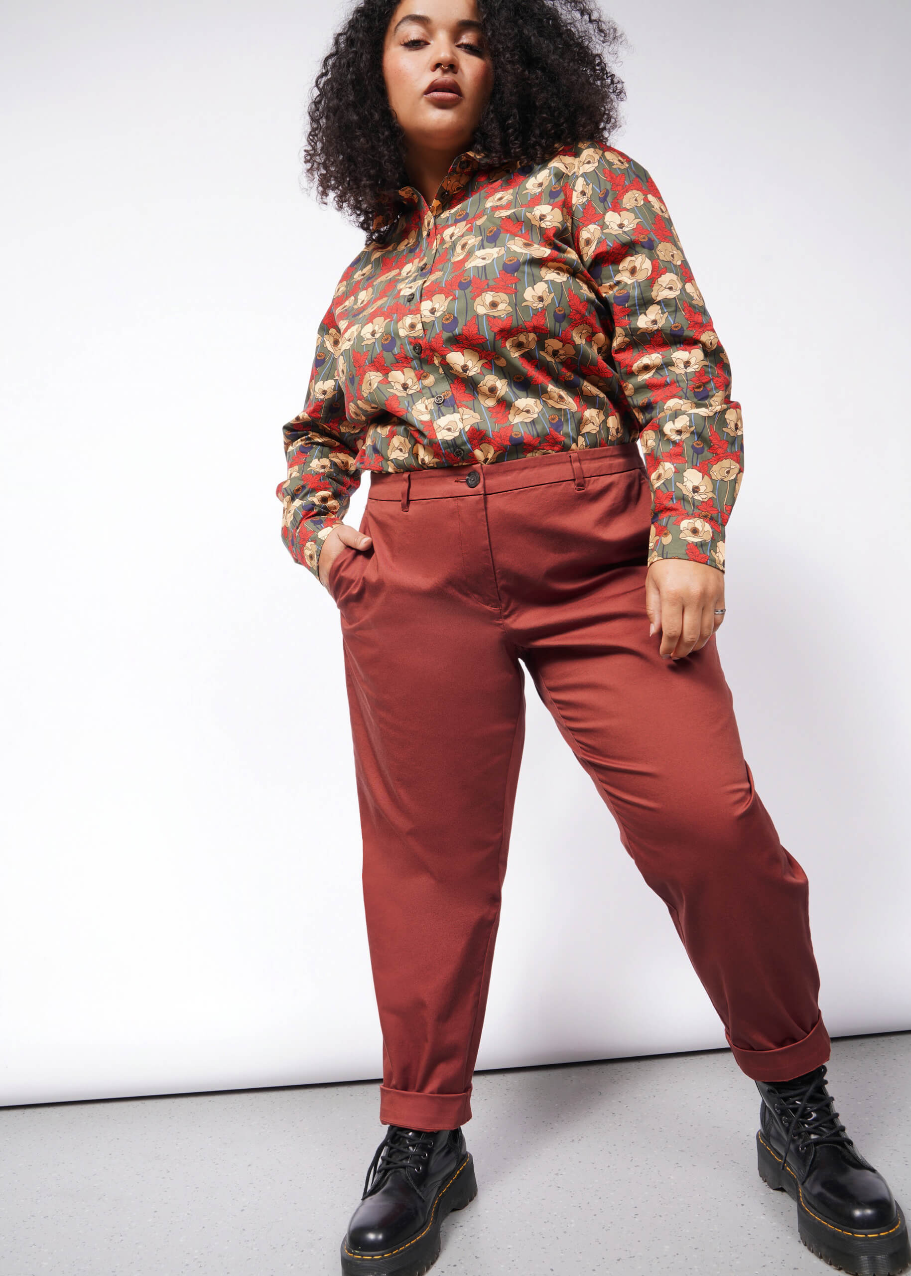 A person with curly hair wears a colorful, patterned shirt and The Essential Trouser in rust color with a relaxed mid-rise fit, paired with black boots. This confident ensemble stands out against a plain white background.