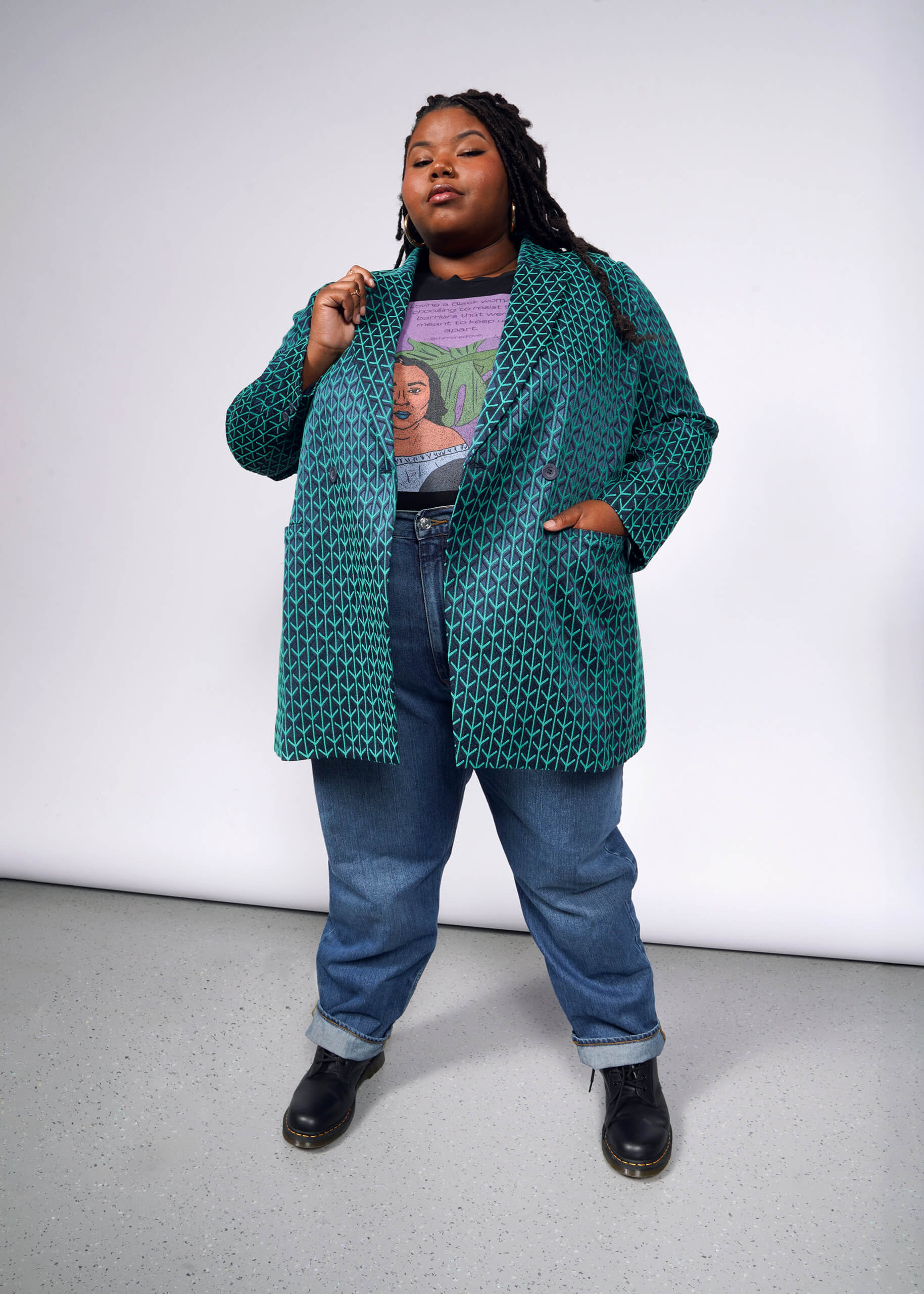 Wearing The Empower Jacquard Double Breasted Blazer—a teal, patterned coat—over a graphic tee with blue jeans and black boots, they stand confidently. Their long braided hair completes the look while one hand is in a pocket and the other adjusts the collar against a plain background.