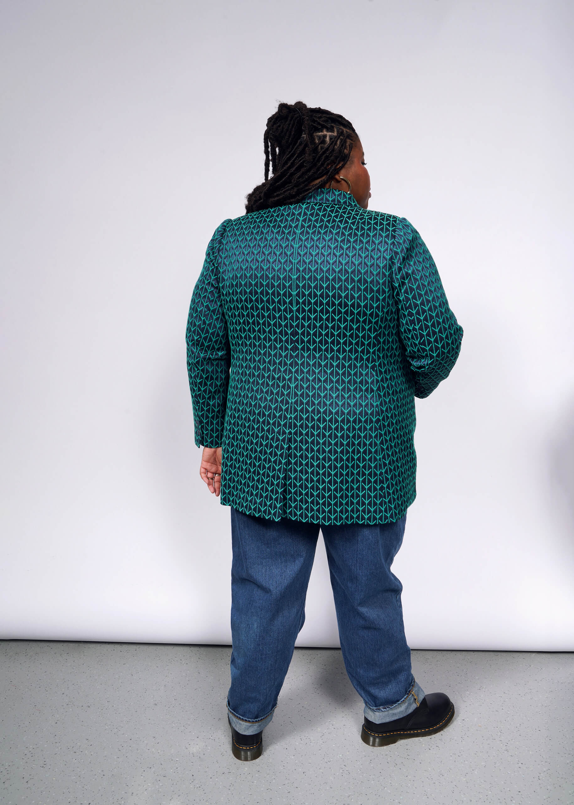 A person with braided hair stands with their back to the camera, showcasing The Empower Jacquard Double Breasted Blazer in teal and black, hinting at a tailored look. Complemented by blue jeans and black shoes, they stand against a plain, light-colored wall and floor.