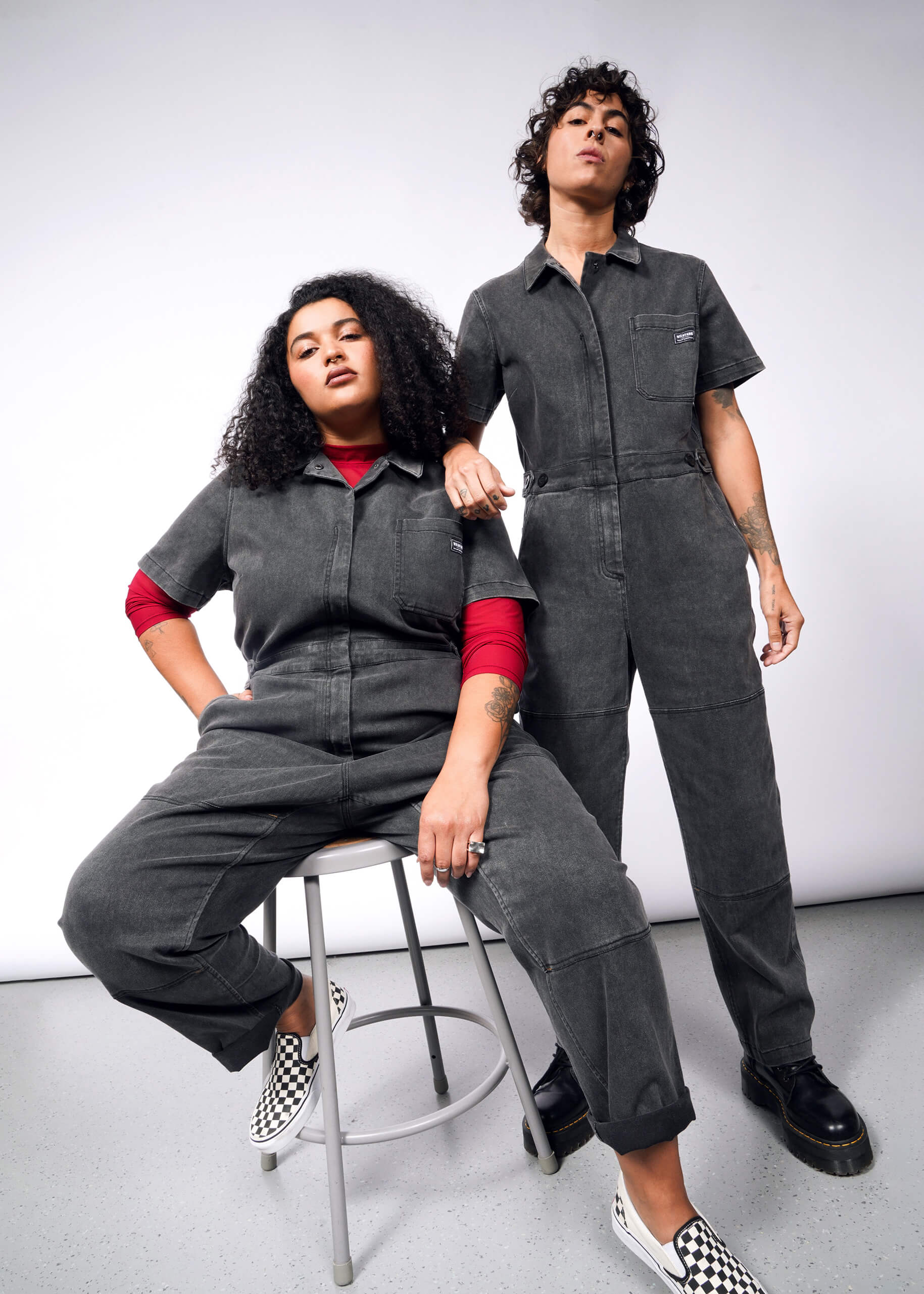 Two individuals confidently model The Essential Denim High Waisted Coverall against a plain backdrop. One sits on a stool with curly hair and checkered shoes, while the other stands with short hair; both gaze at the camera.