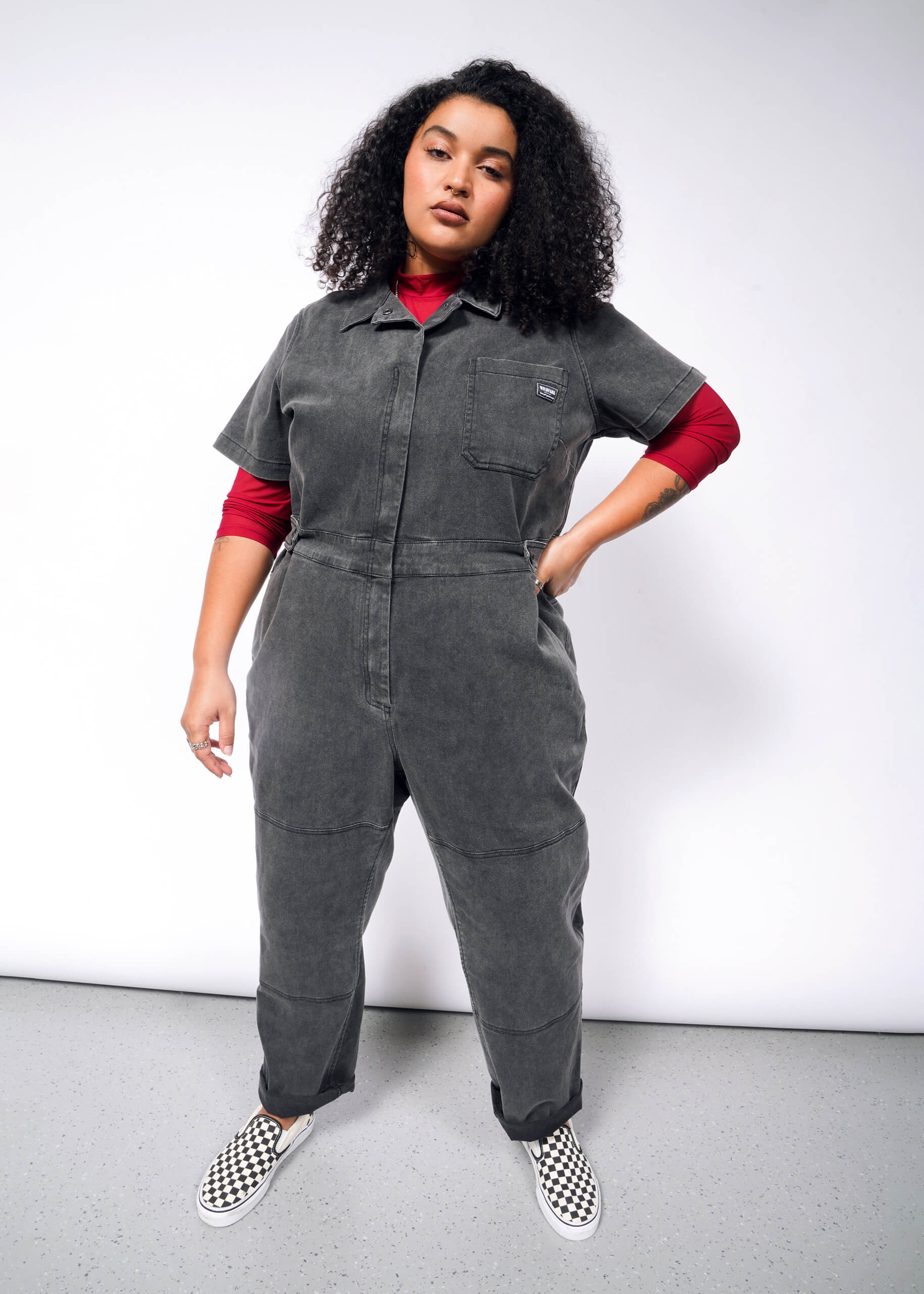 A person with curly hair confidently wears The Essential Denim High Waisted Coverall in gray over a red turtleneck, paired with checkerboard-patterned slip-on shoes, against a plain white backdrop.