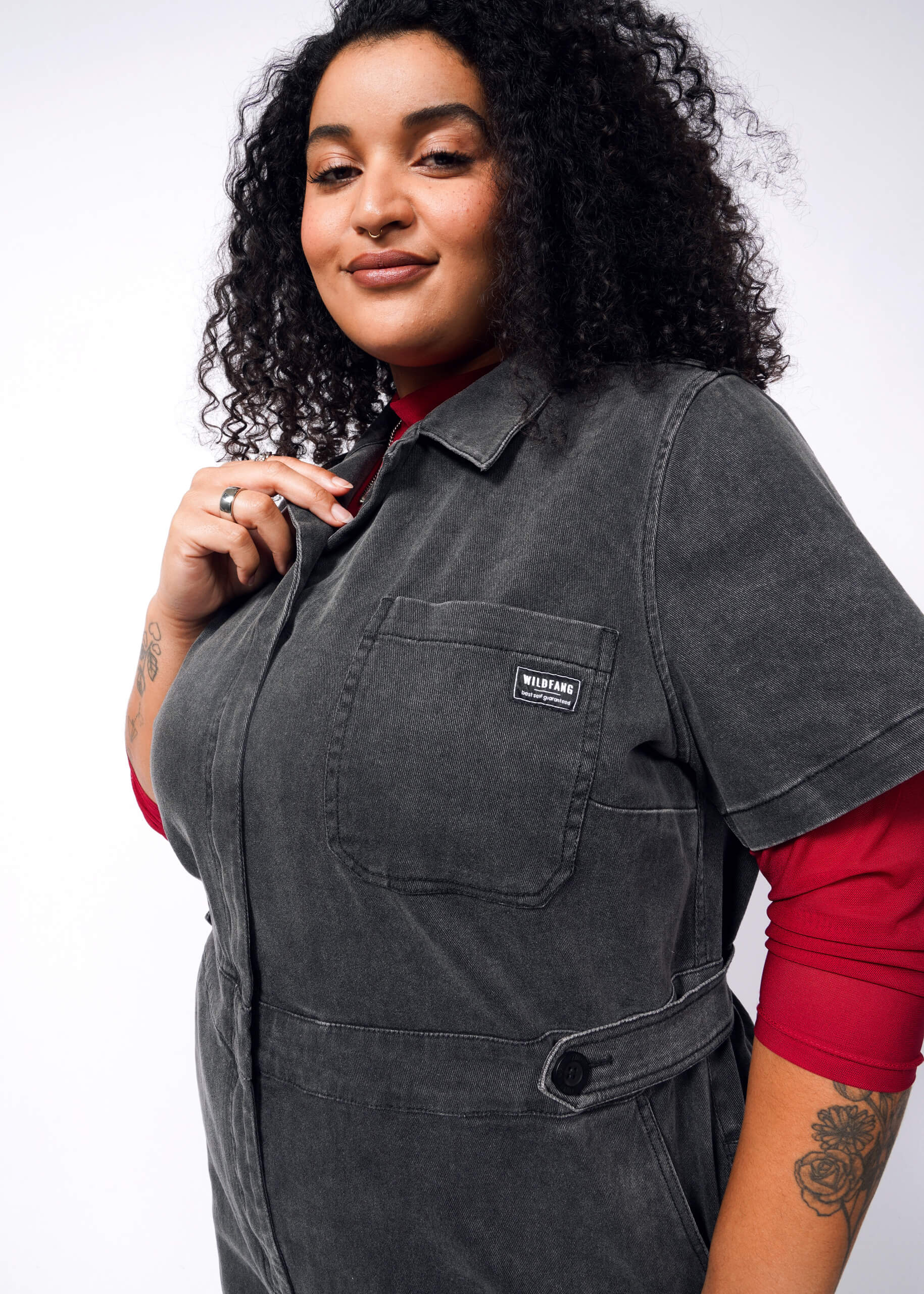 A person with curly hair smiles, wearing The Essential Denim High Waisted Coverall in dark gray over a red top, showcasing arm tattoos while holding the jumpsuits lapel against a plain white background.