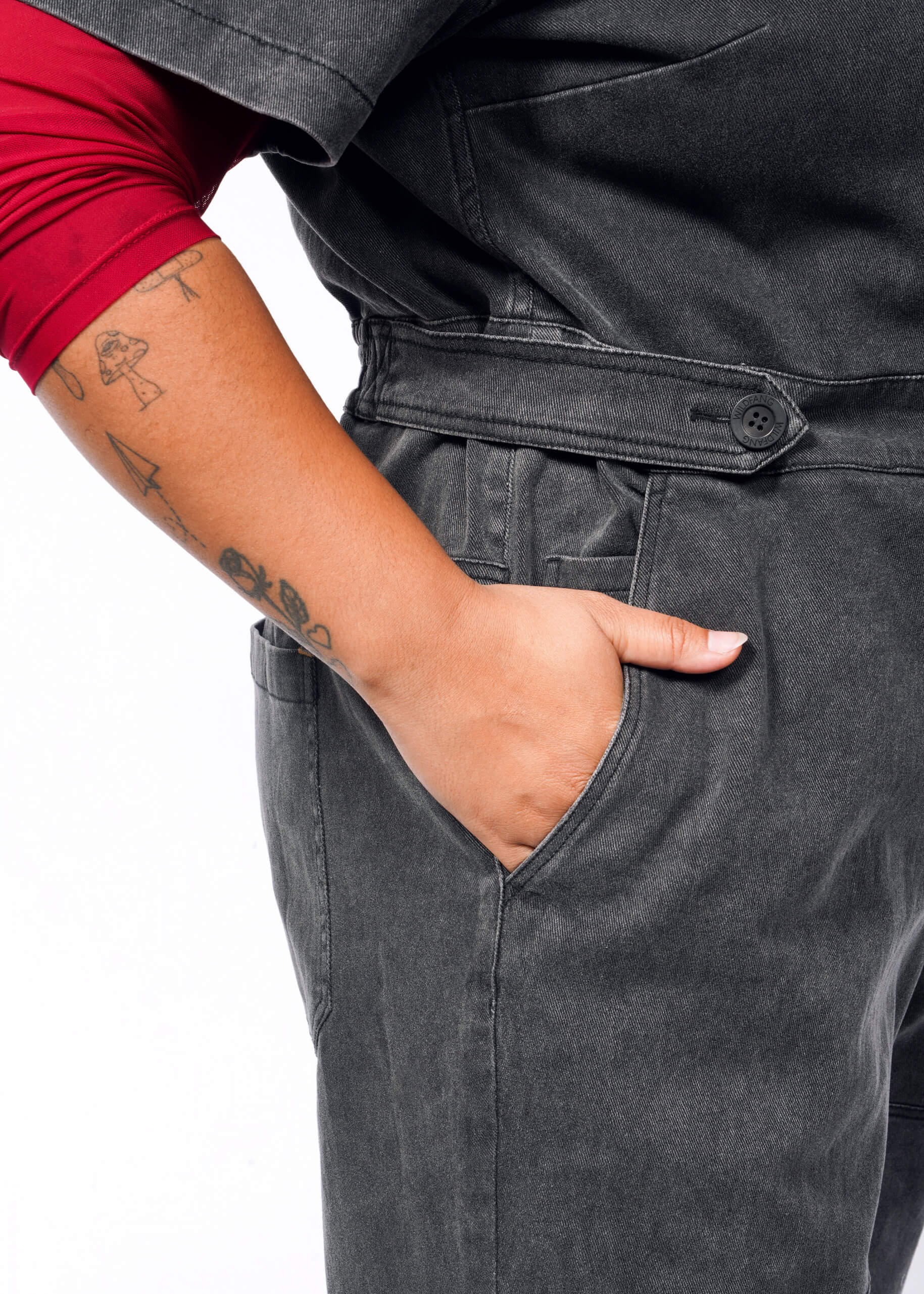 A person with tattoos on their forearm wears The Essential Denim High Waisted Coverall in dark gray. It features an adjustable waist and deep pockets, with one hand casually tucked in. A red sleeve peeks out as they stand against a plain white background.