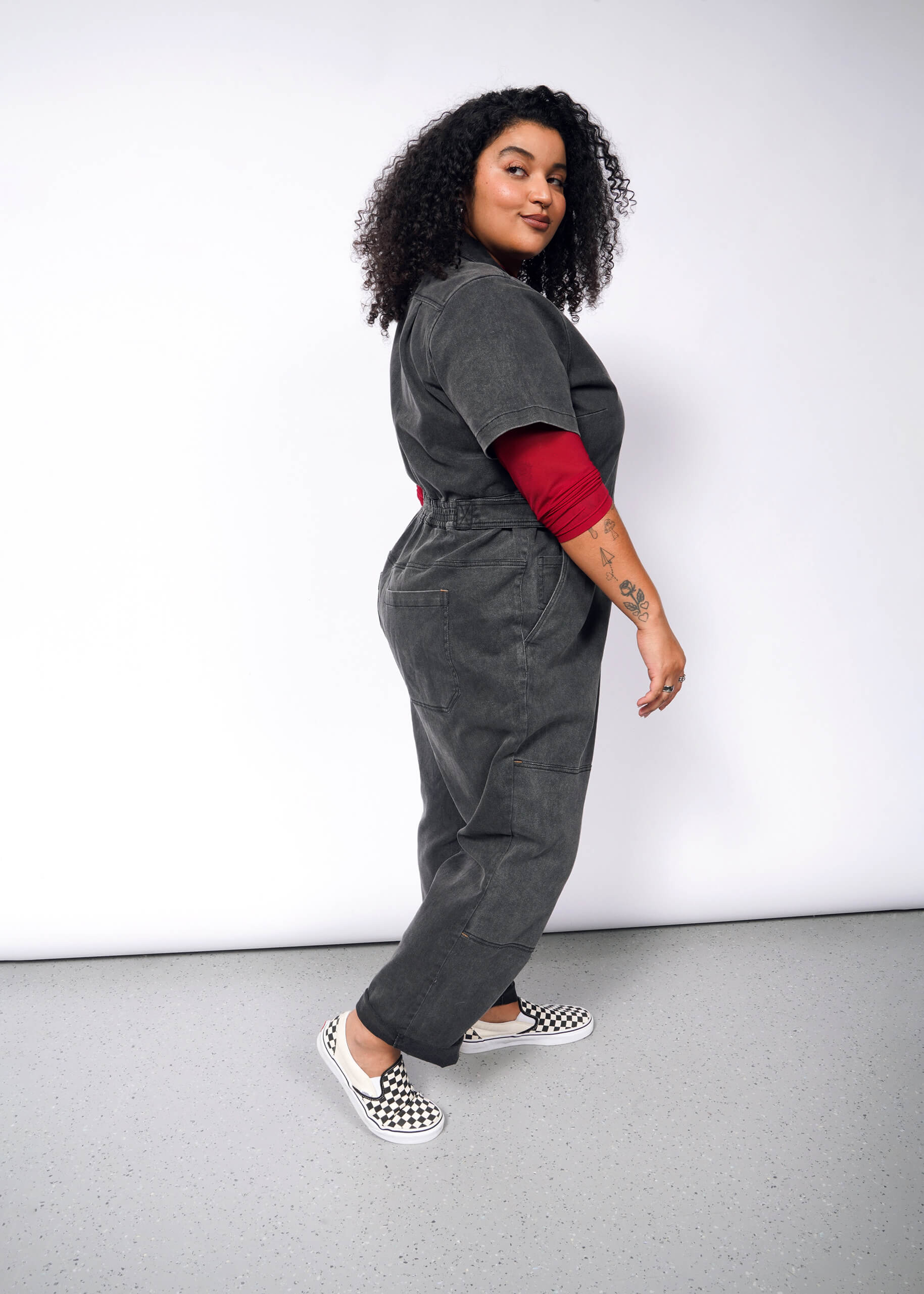 With tattoos visible on their forearm, a person with curly hair poses confidently in The Essential Denim High Waisted Coverall over a red long-sleeve shirt, paired with checkered shoes against a plain light gray wall.