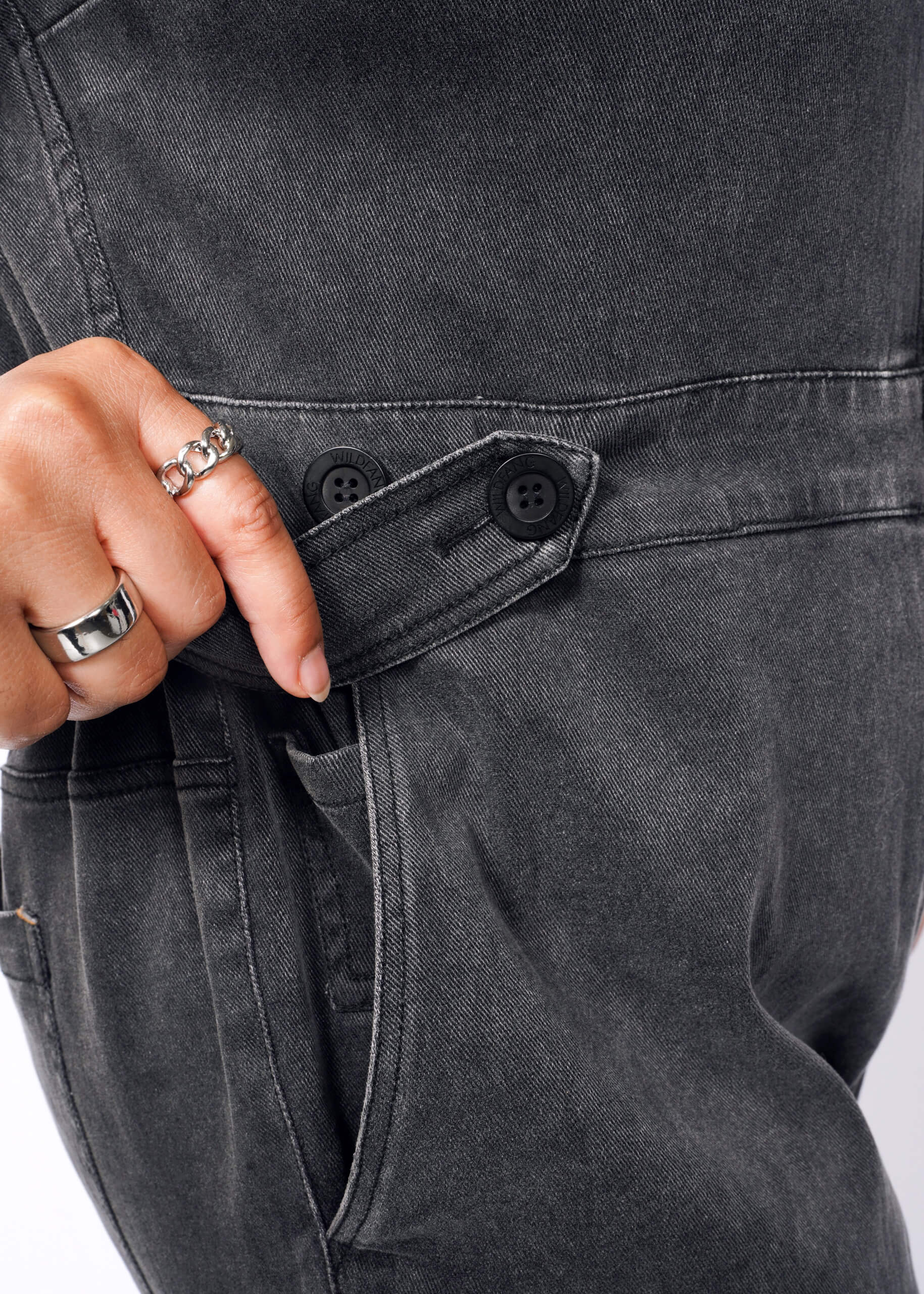 A person wearing silver rings adjusts the buttoned strap on The Essential Denim High Waisted Coverall, in dark gray. The close-up highlights their hand and the coveralls adjustable waist area, focusing on texture and stitching.