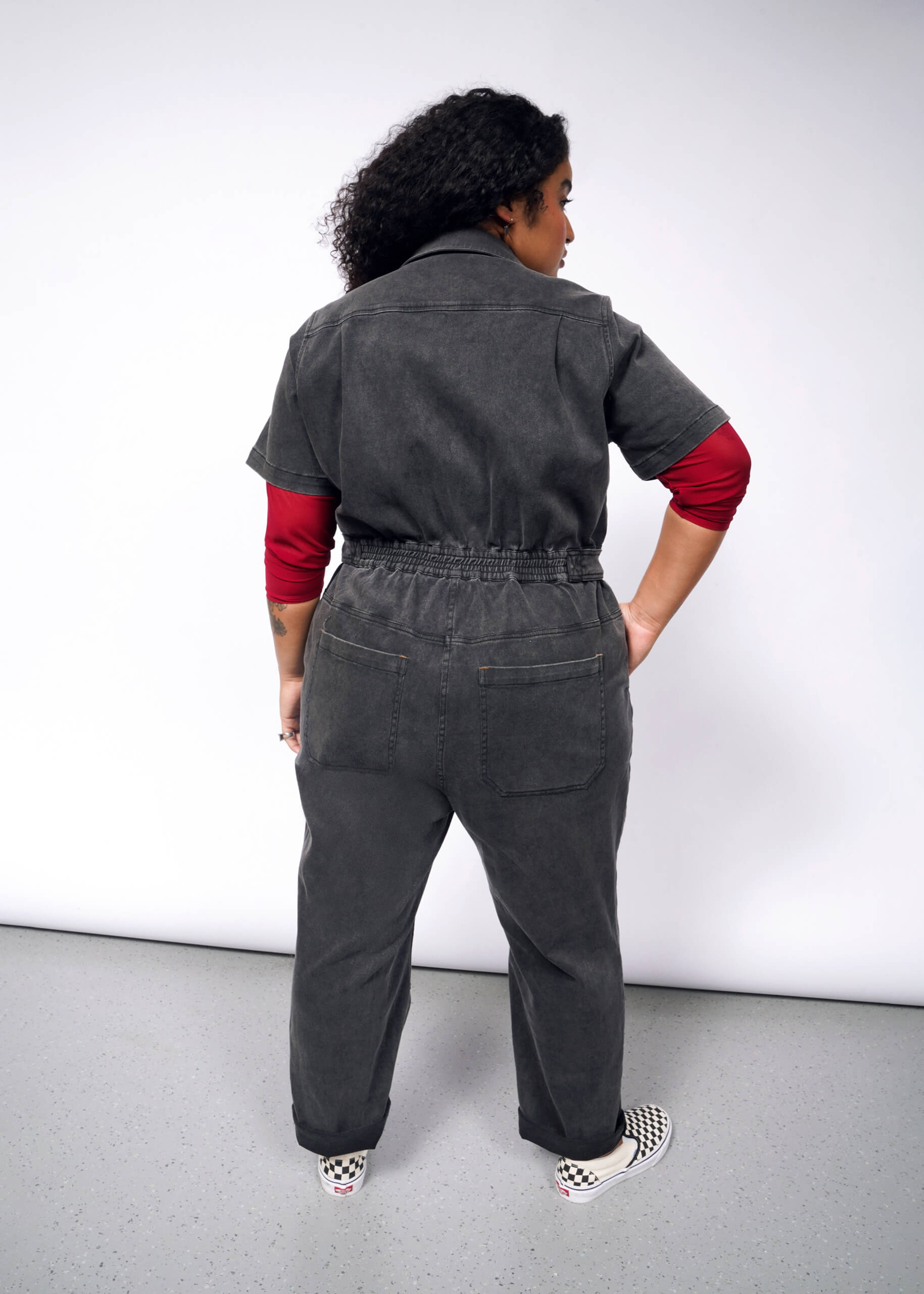 A person with curly hair, back to the camera, wears The Essential Denim High Waisted Coverall—a gray jumpsuit with deep pockets—over a red long-sleeve shirt and checkered shoes, hands on hips against a plain white background.