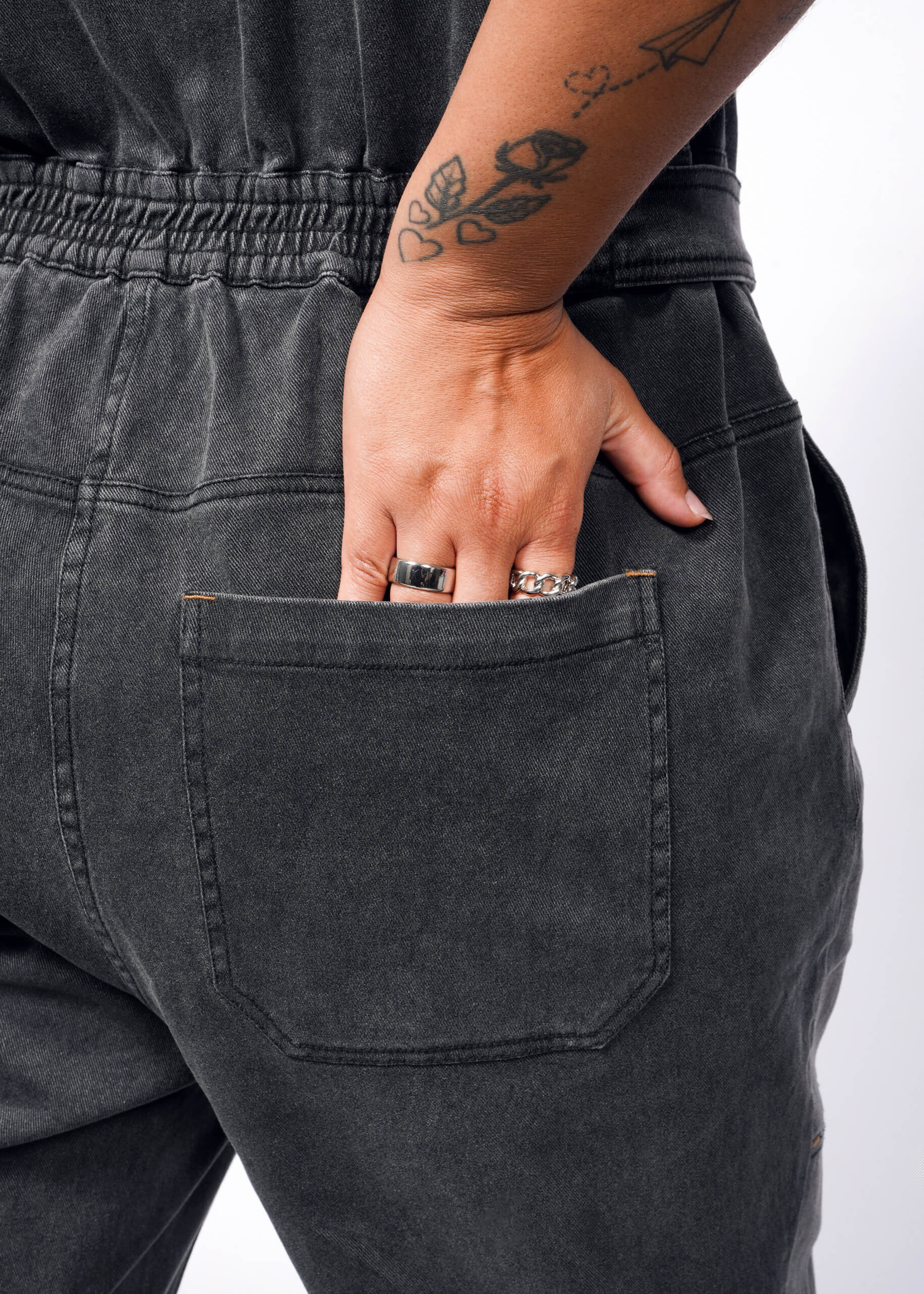 A tattooed person sporting The Essential Denim High Waisted Coverall in dark gray places their left hand, adorned with several rings, into a deep pocket with a gathered waistband.