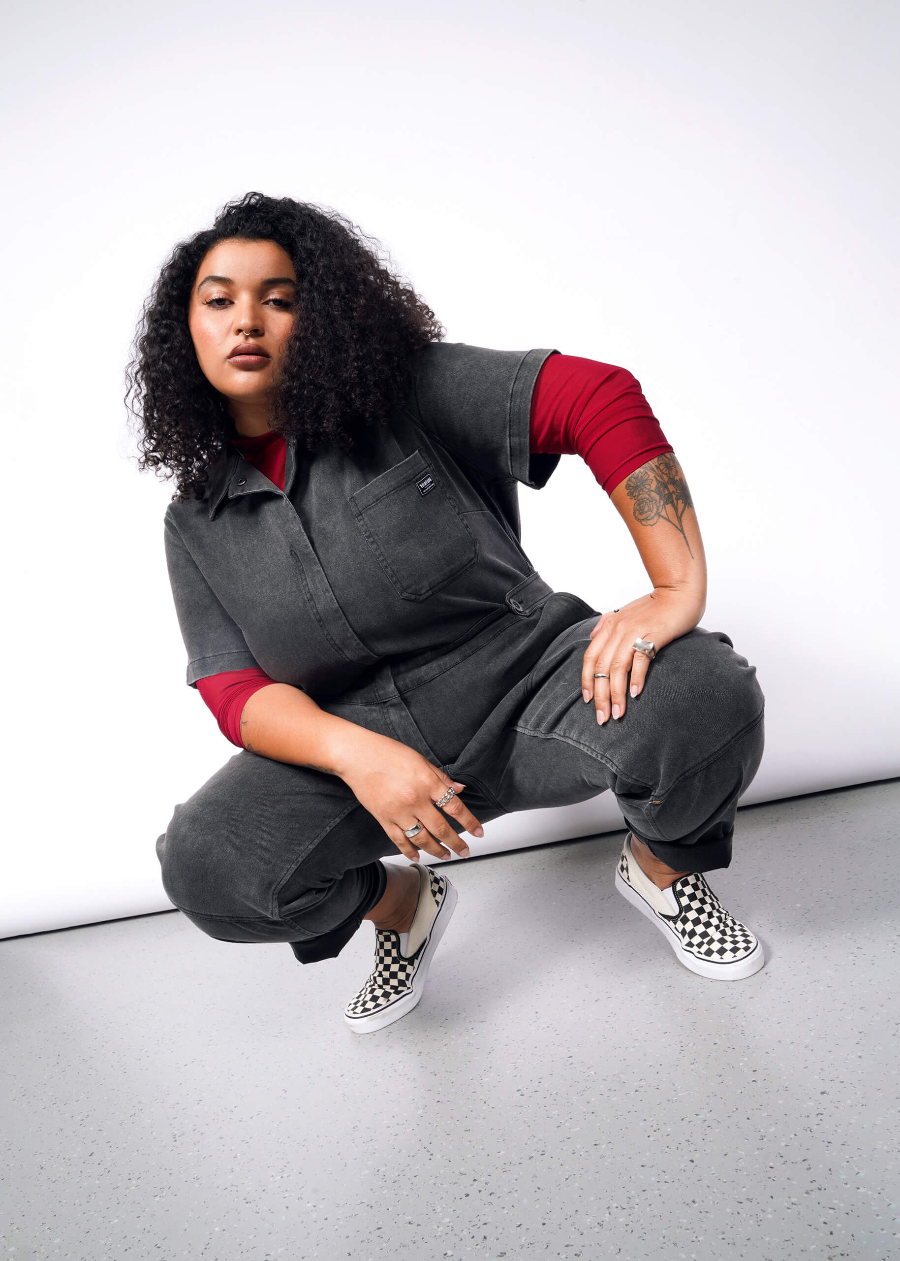 A person with curly hair confidently squats, wearing The Essential Denim High Waisted Coverall and a red long-sleeve shirt. Checkered slip-on shoes complement the look, revealing tattoos on their arm as hands rest on knees. A plain, light-colored background completes the scene.