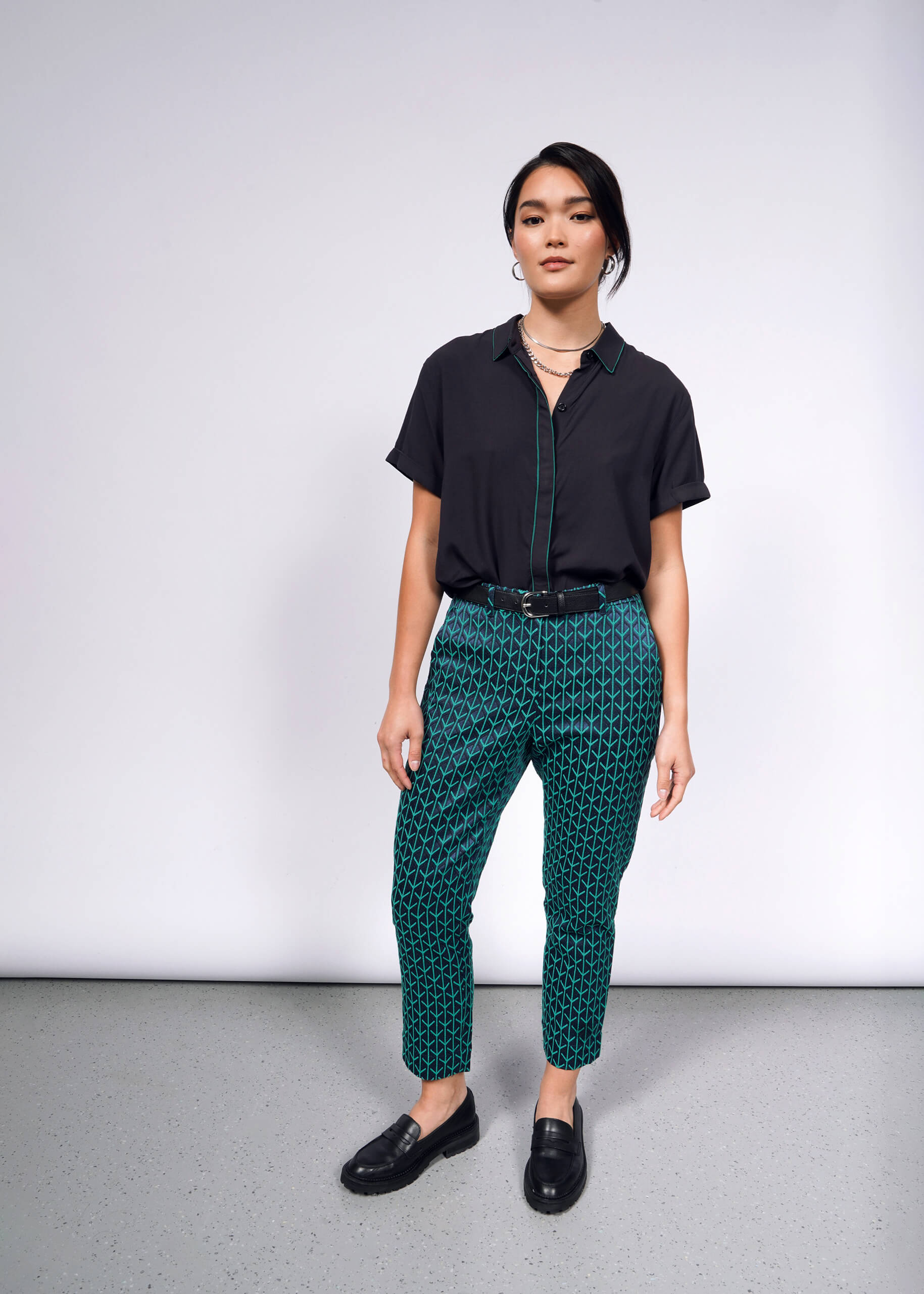 A person stands against a plain backdrop wearing a dark short-sleeved shirt, The Empower Jacquard Slim Crop Pant in green with a high-rise fit, and black loafers, accessorized with layered necklaces.