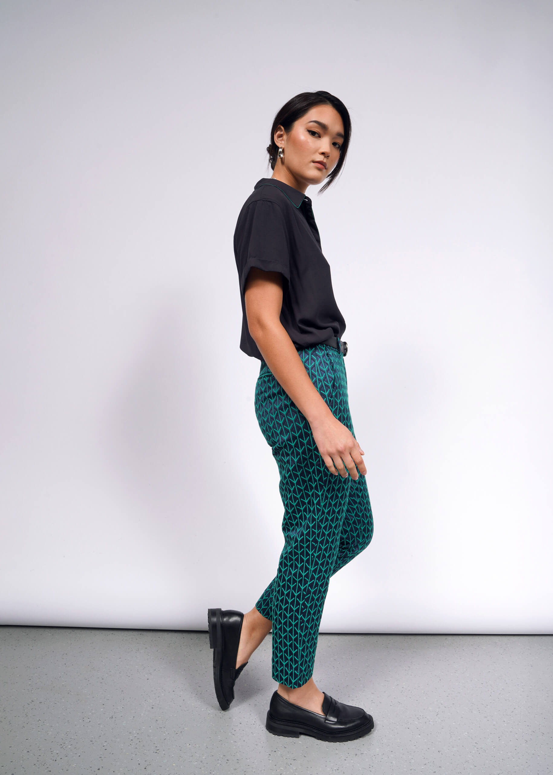 A person in a dark blouse and The Empower Jacquard Slim Crop Pant, featuring a green jacquard pattern, strolls sideways against a plain background. They have short hair and wear sleek black loafers.