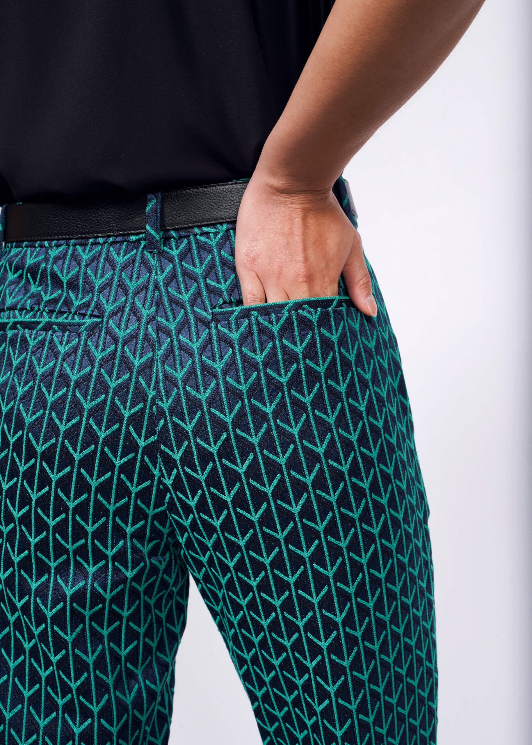 A person wearing The Empower Jacquard Slim Crop Pant, a vibrant cropped pant featuring a green and blue geometric arrow pattern on jacquard fabric, sports a black belt and casually places their hand in their pocket against a plain white background.