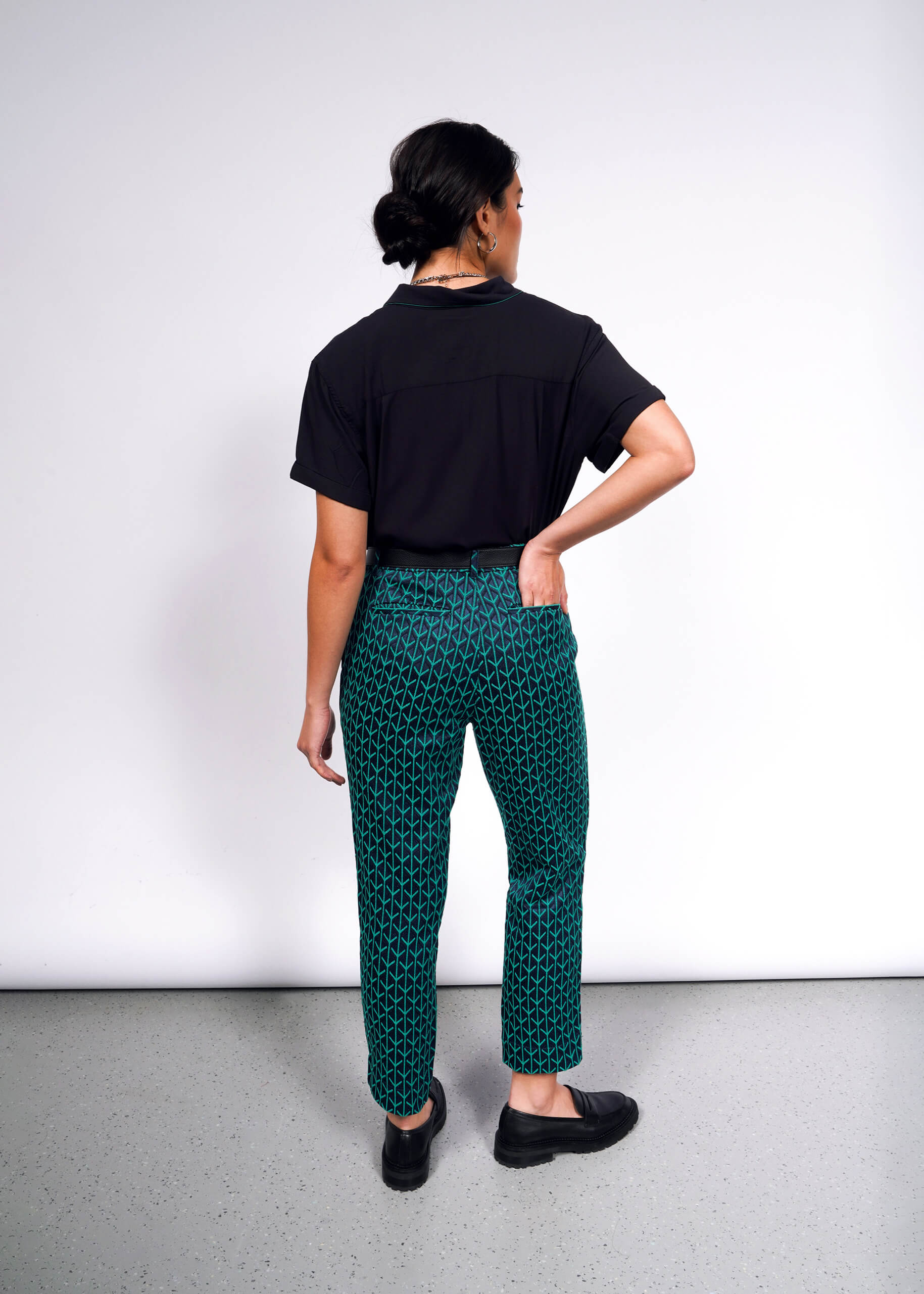 A person stands with their back to the camera on a gray floor against a white wall, wearing a black shirt and The Empower Jacquard Slim Crop Pant in green. Their hand rests on their hip.