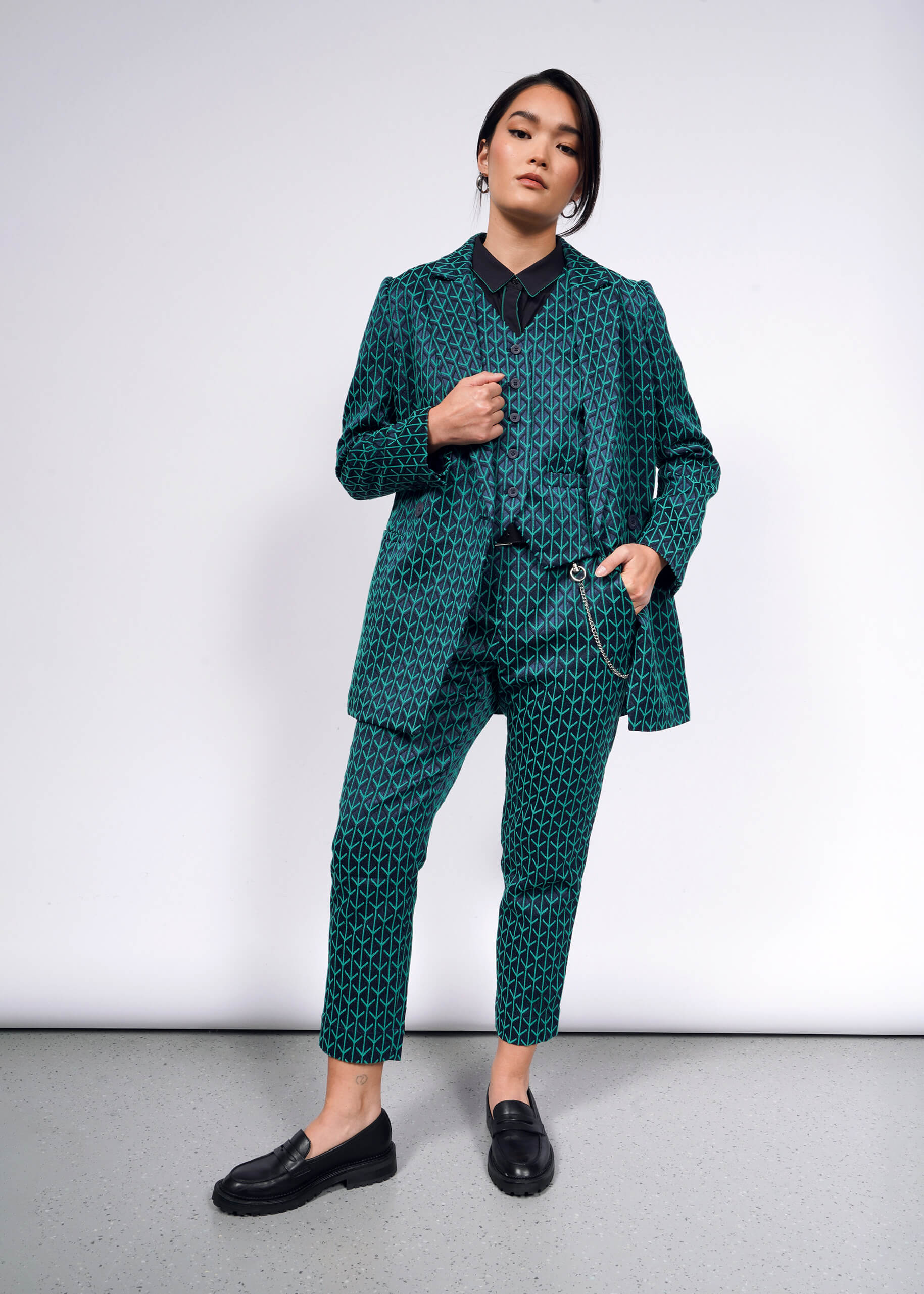 A person stands confidently against a plain background, showcasing The Empower Jacquard Double Breasted Blazer in teal. With their hands in pockets and sporting black loafers, their hair is tied back, complementing the ensemble with a neutral expression.