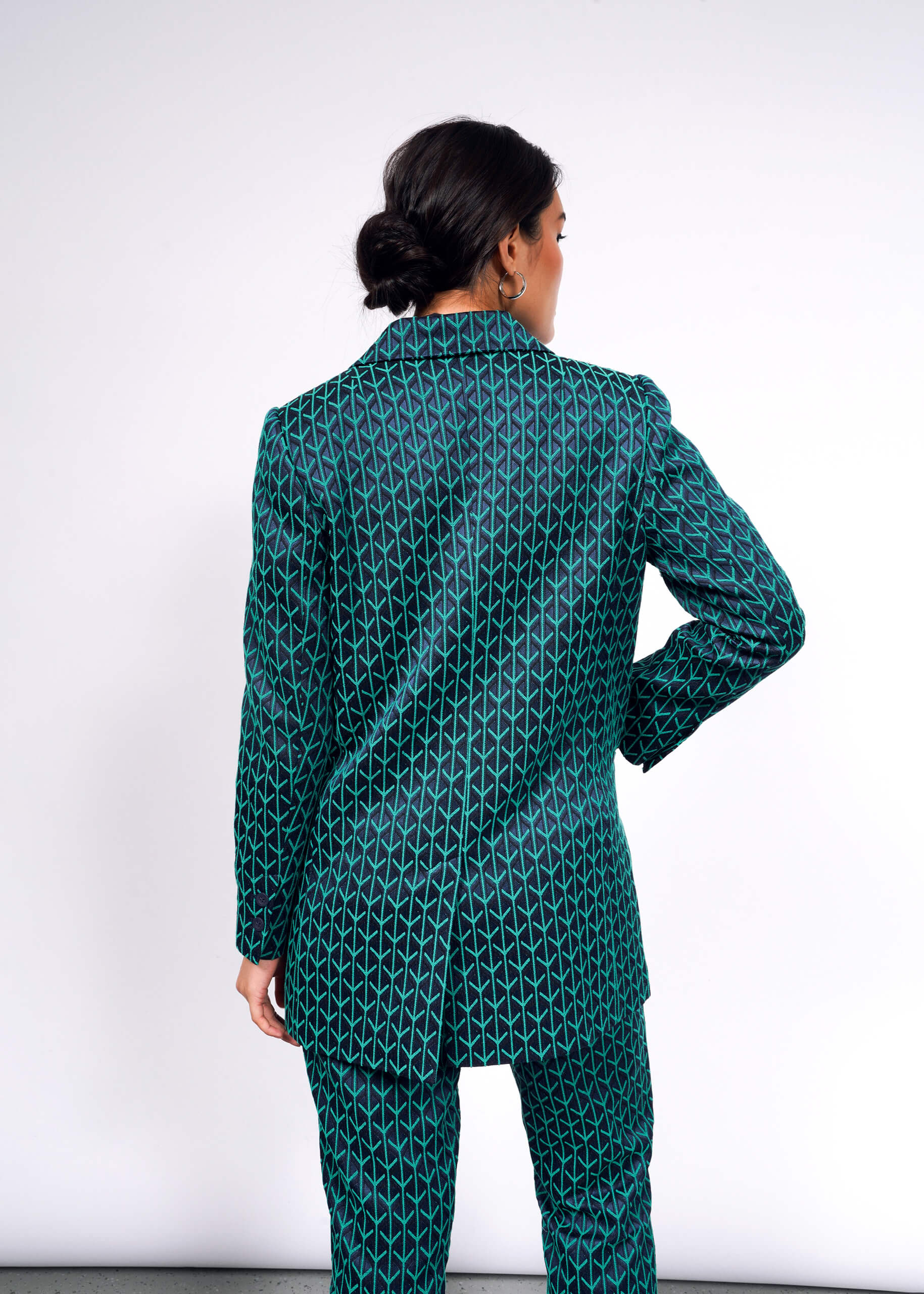 A person with dark hair styled in an updo is facing away while wearing The Empower Jacquard Double Breasted Blazer, a green suit featuring a geometric design on a white background, highlighting its tailored fit.