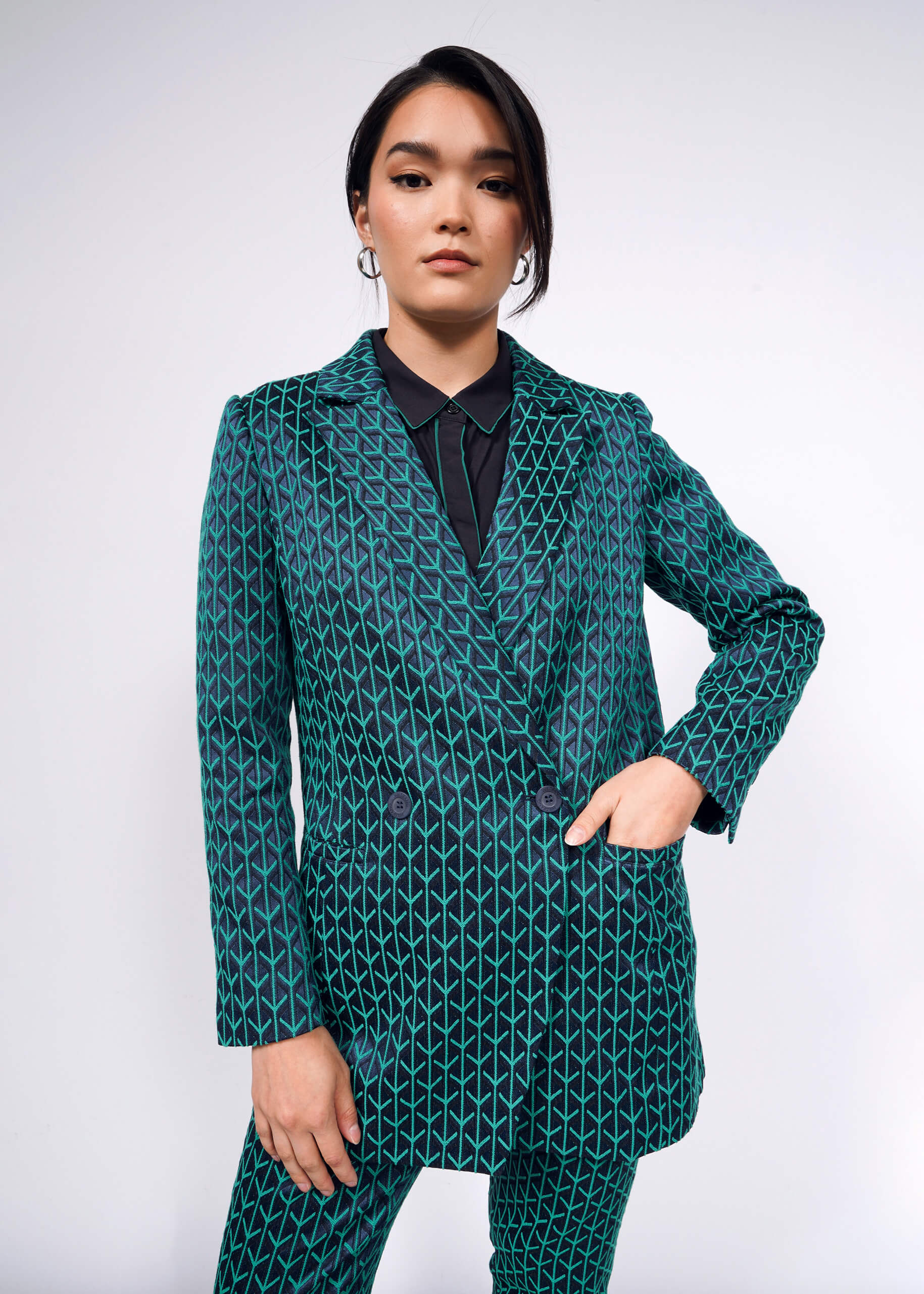 Standing confidently, a person wears The Empower Jacquard Double Breasted Blazer in teal with a geometric pattern over a black shirt, styled with hoop earrings and sleek hair. The woven jacquard fabric adds sophistication against the plain, light-colored background.