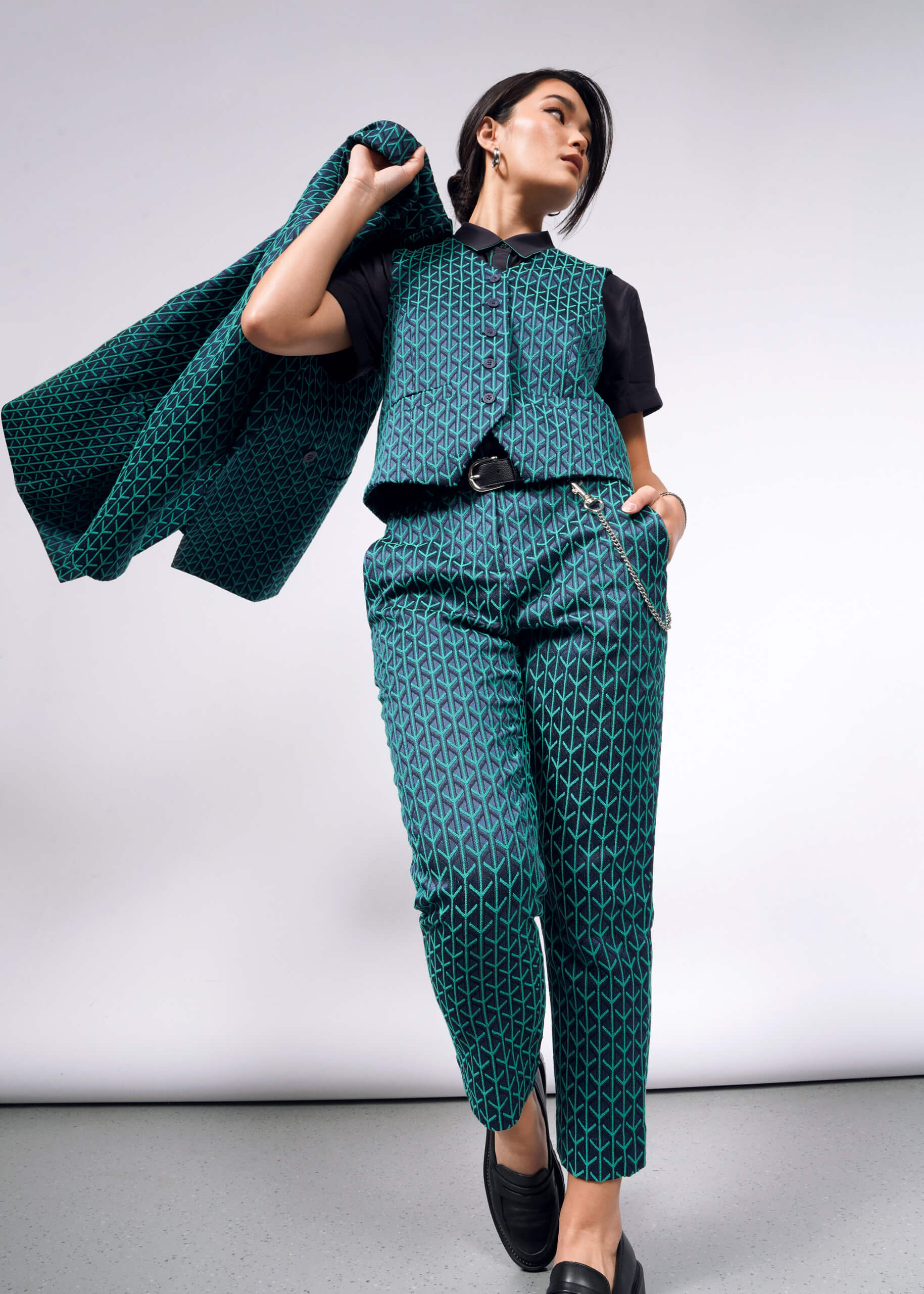 Dressed in The Empower Jacquard Slim Crop Pant, a person stands confidently against a gray backdrop. They carry the matching geometric-patterned green jacket over their shoulder, wearing a black shirt and loafers for an elegant touch.