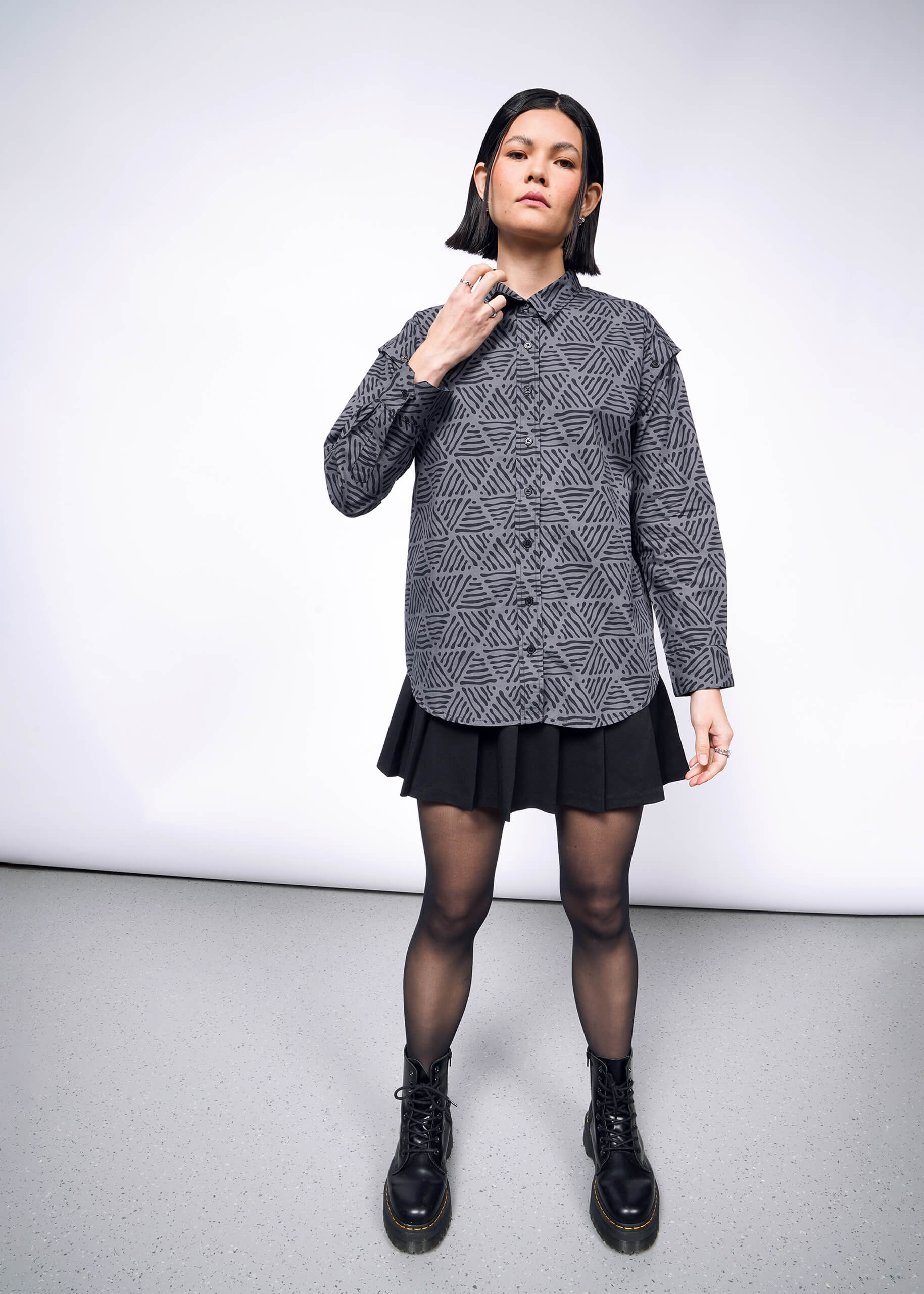 A person with short black hair models The Essential Convertible Sleeve Button Up, a versatile patterned gray shirt, paired with a pleated black skirt. They complete the look with sheer black tights and boots, striking a pose against a plain backdrop while their right hand touches their collar.