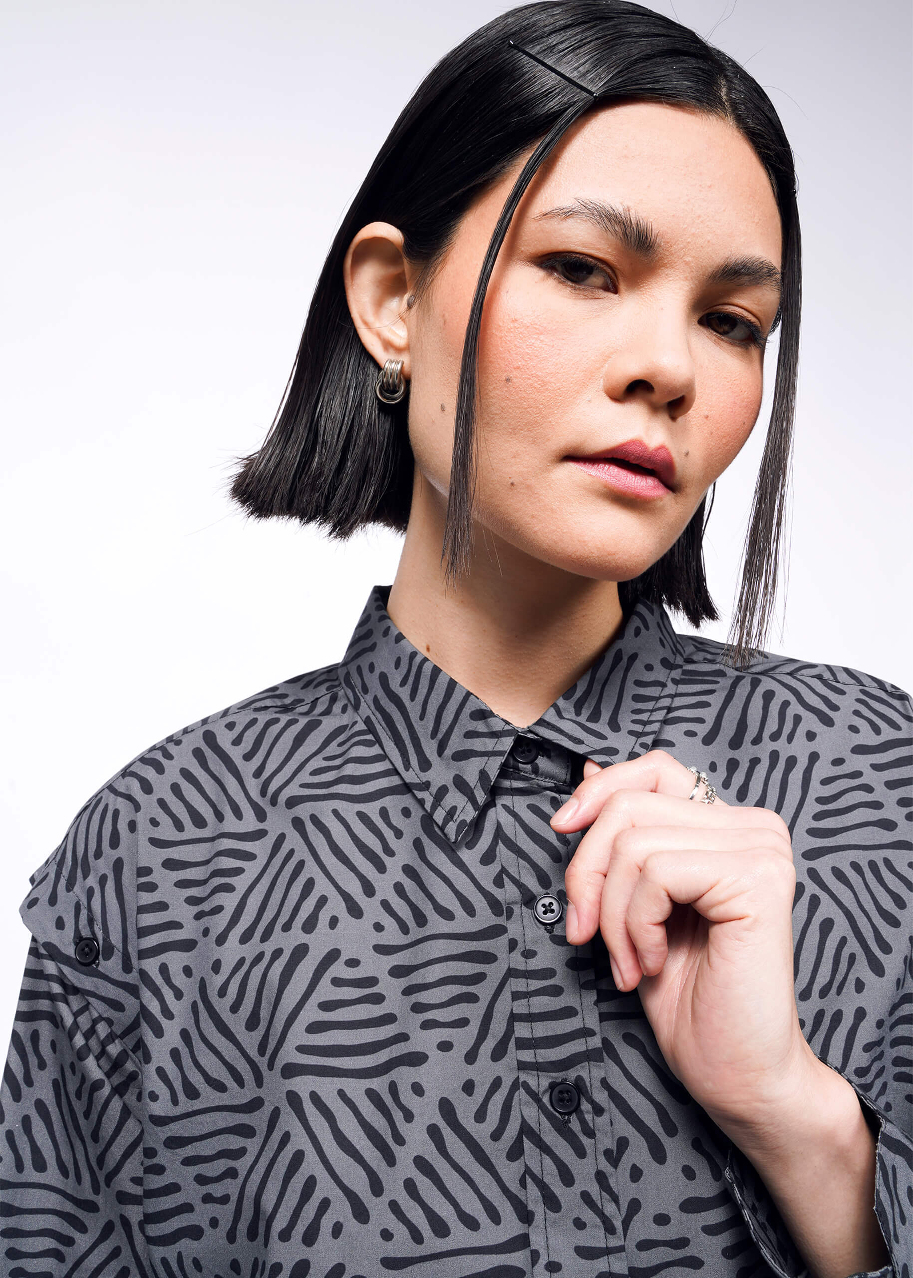 A person with short black hair is wearing The Essential Convertible Sleeve Button Up in gray cotton, styled with silver earrings. They look at the camera neutrally, one hand near their collar. The background is plain white.