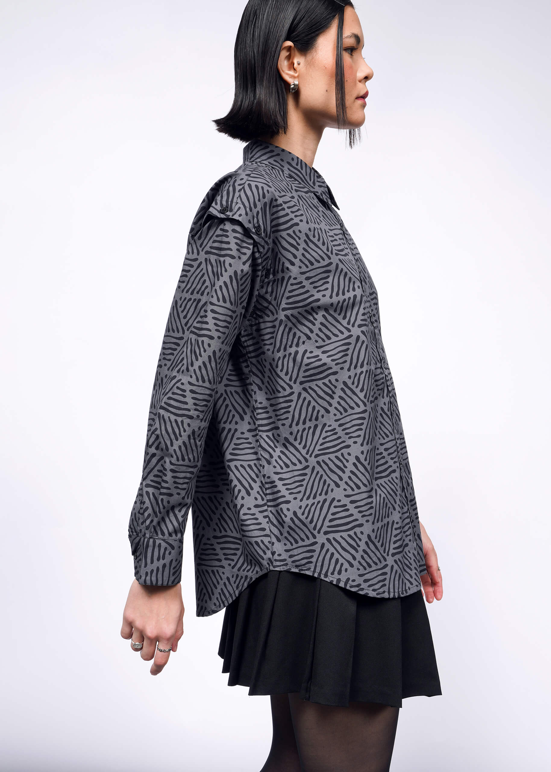 A person with shoulder-length black hair is shown in profile wearing The Essential Convertible Sleeve Button Up—a patterned gray cotton shirt paired with a black skirt. The plain white background highlights the geometric design and removable sleeves of this versatile piece.