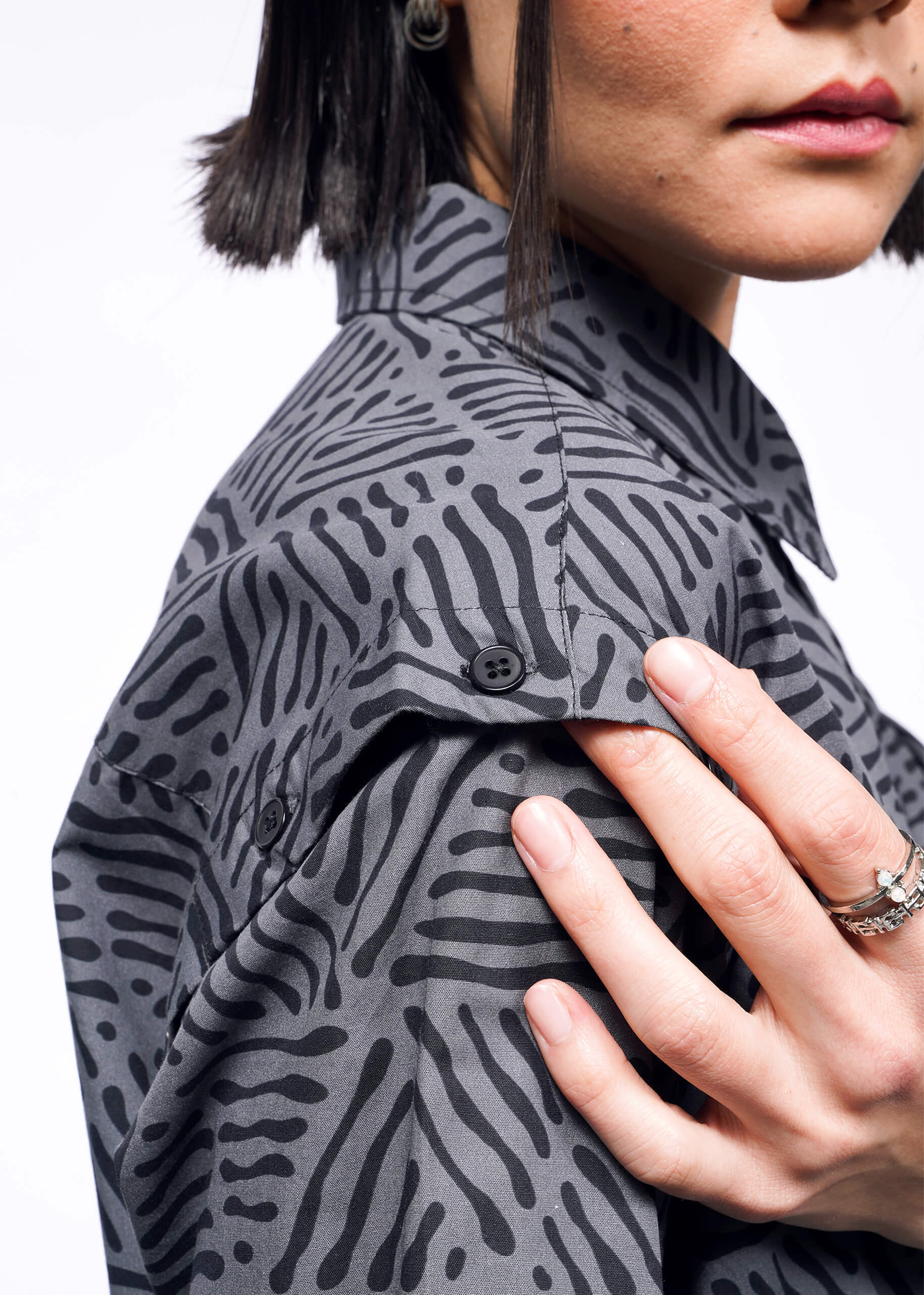 A person with short, dark hair wears The Essential Convertible Sleeve Button Up, showcasing its black abstract pattern against a white background. Adjusting the cuff with their left hand, adorned with a ring, they highlight the shirts elegance.