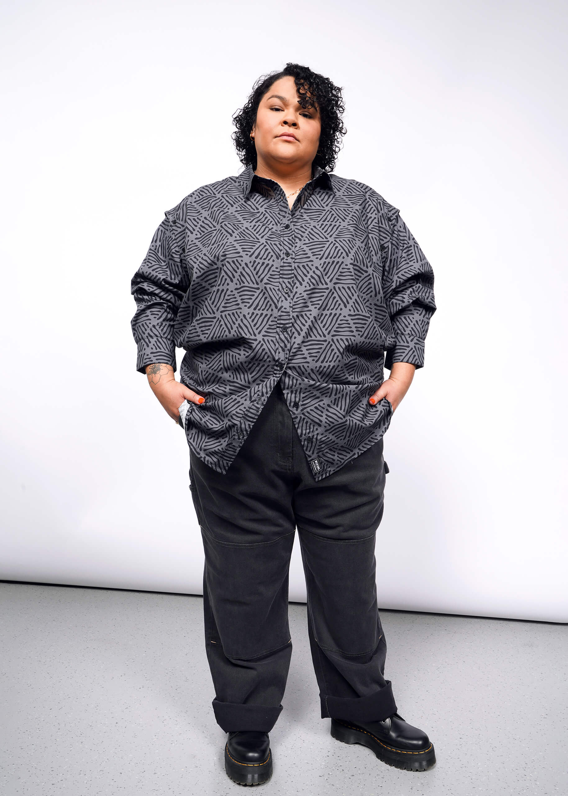 A person with short curly hair stands confidently against a plain background, wearing The Essential Convertible Sleeve Button Up featuring a versatile geometric pattern, paired with dark pants, hands in pockets.