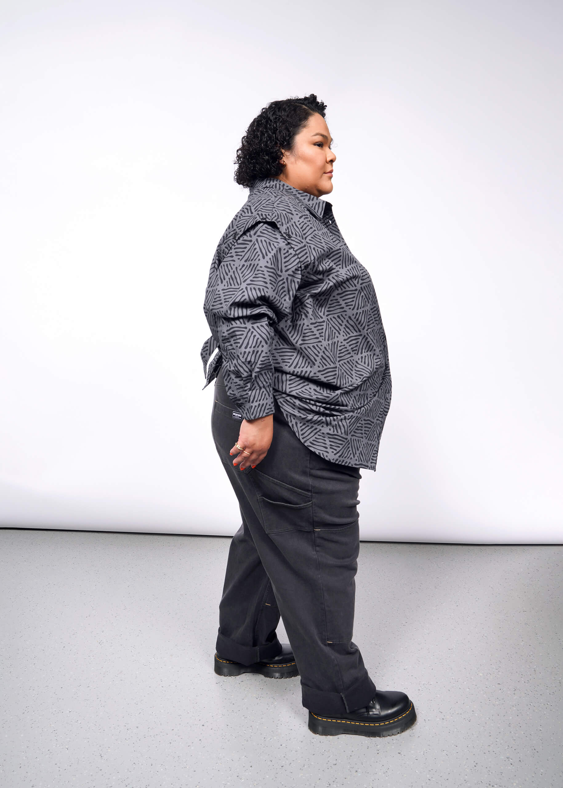 Against a plain backdrop, a person with curly hair stands in profile wearing The Essential Convertible Sleeve Button Up featuring a geometric pattern, paired with dark pants and black shoes. Their expression is neutral as they stand on a light gray floor.