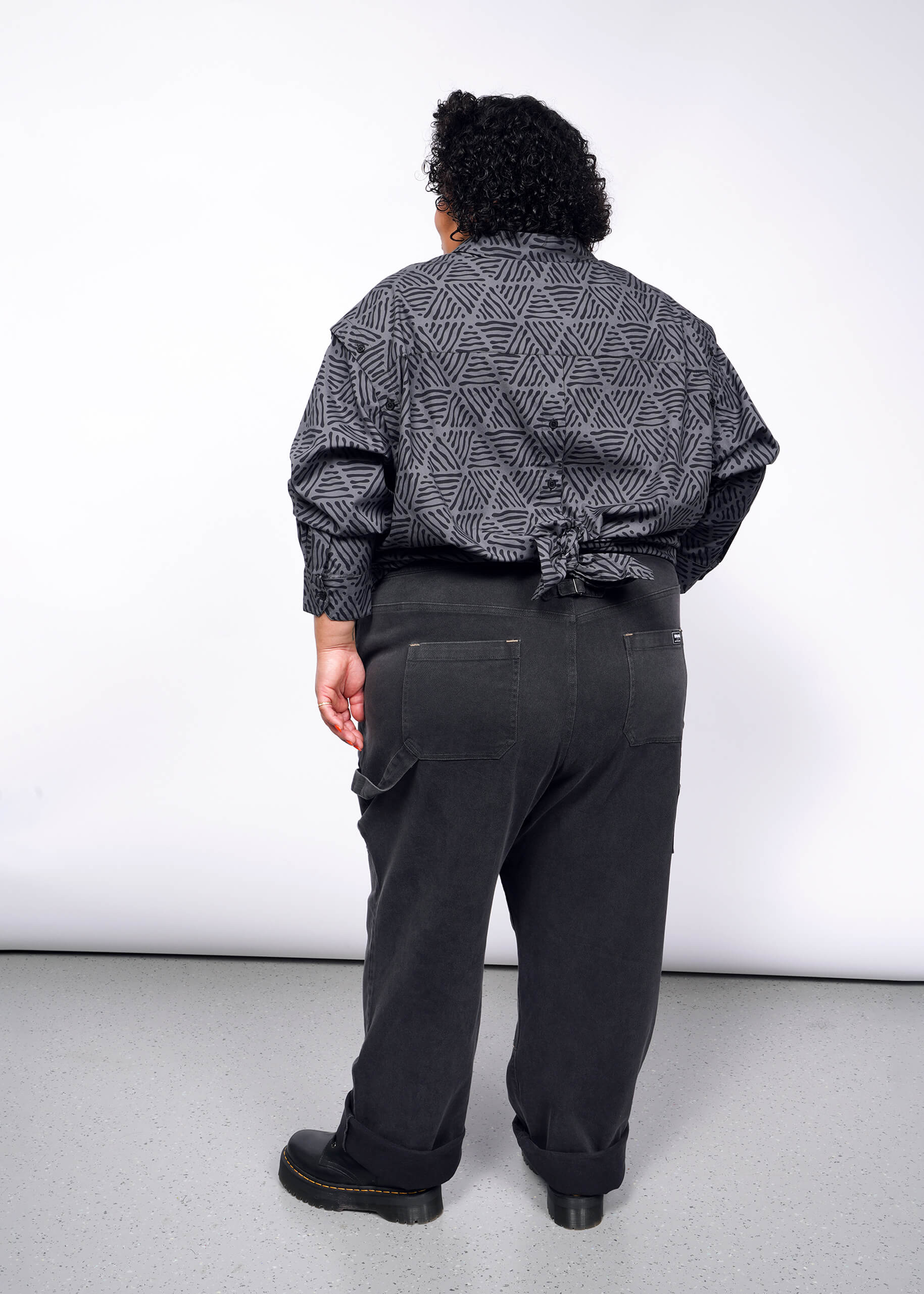 A person faces a plain, light-colored wall, wearing The Essential Convertible Sleeve Button Up, a geometric-patterned gray jacket with removable sleeves tied at the waist, along with dark pants and shoes. They have short curly hair.